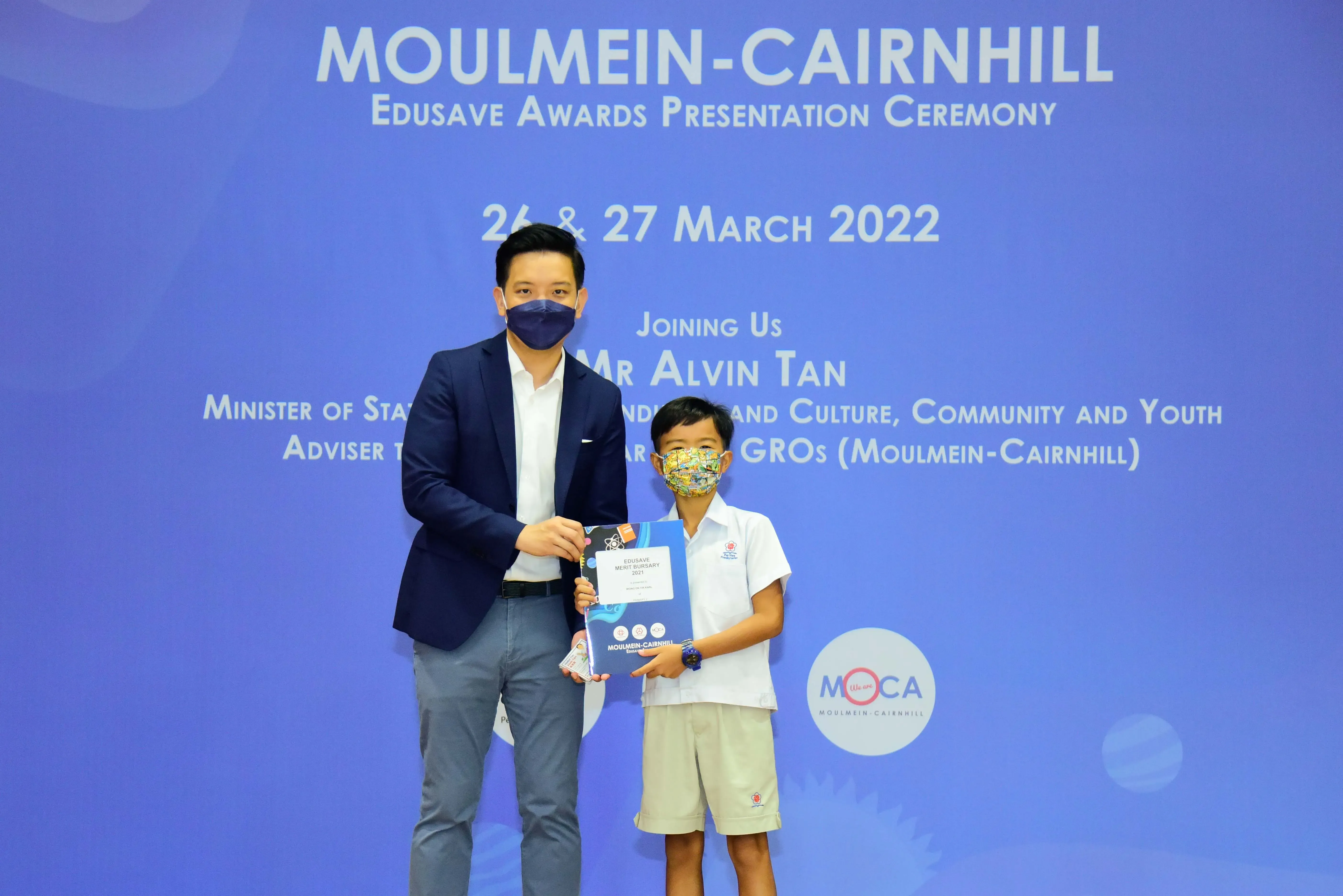 An adult and a young student in masks hold an award at the Moulmein-Cairnhill Edusave Awards Ceremony, March 2022.