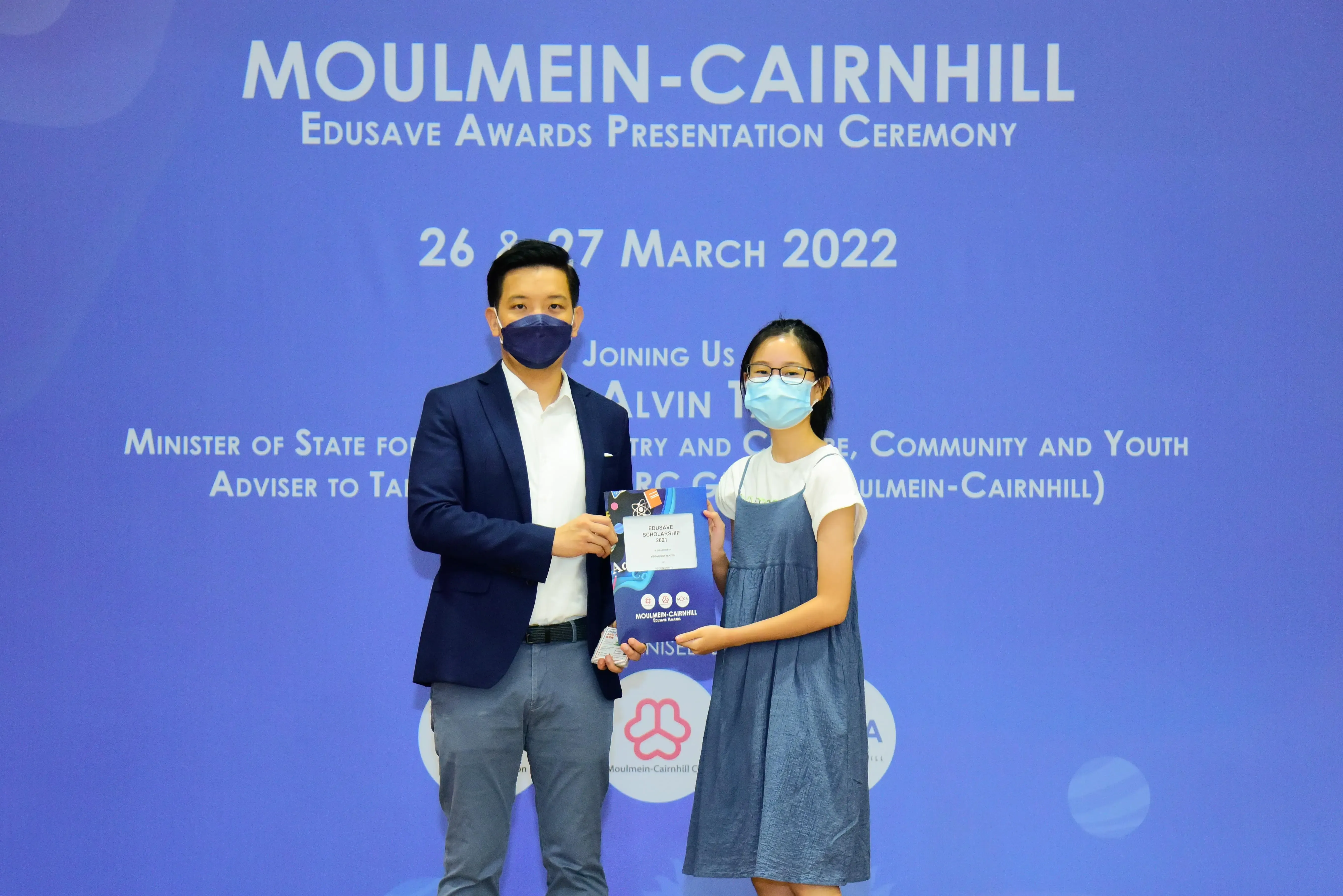 Two masked individuals hold an Edusave Scholarship at the Moulmein-Cairnhill Awards Ceremony, March 2022.