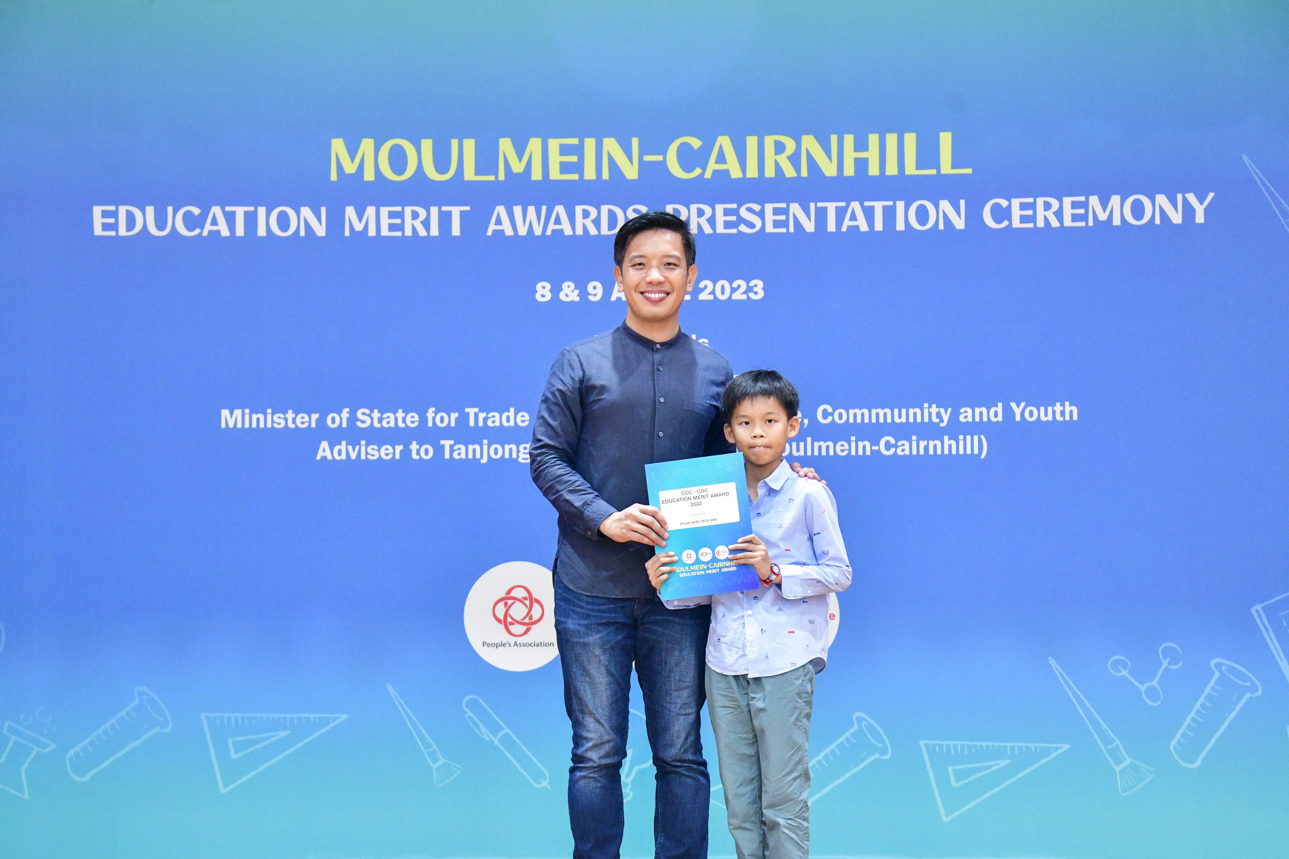 A man and a young boy hold an award certificate in front of a Moulmein-Cairnhill Education Merit Awards 2023 banner.
