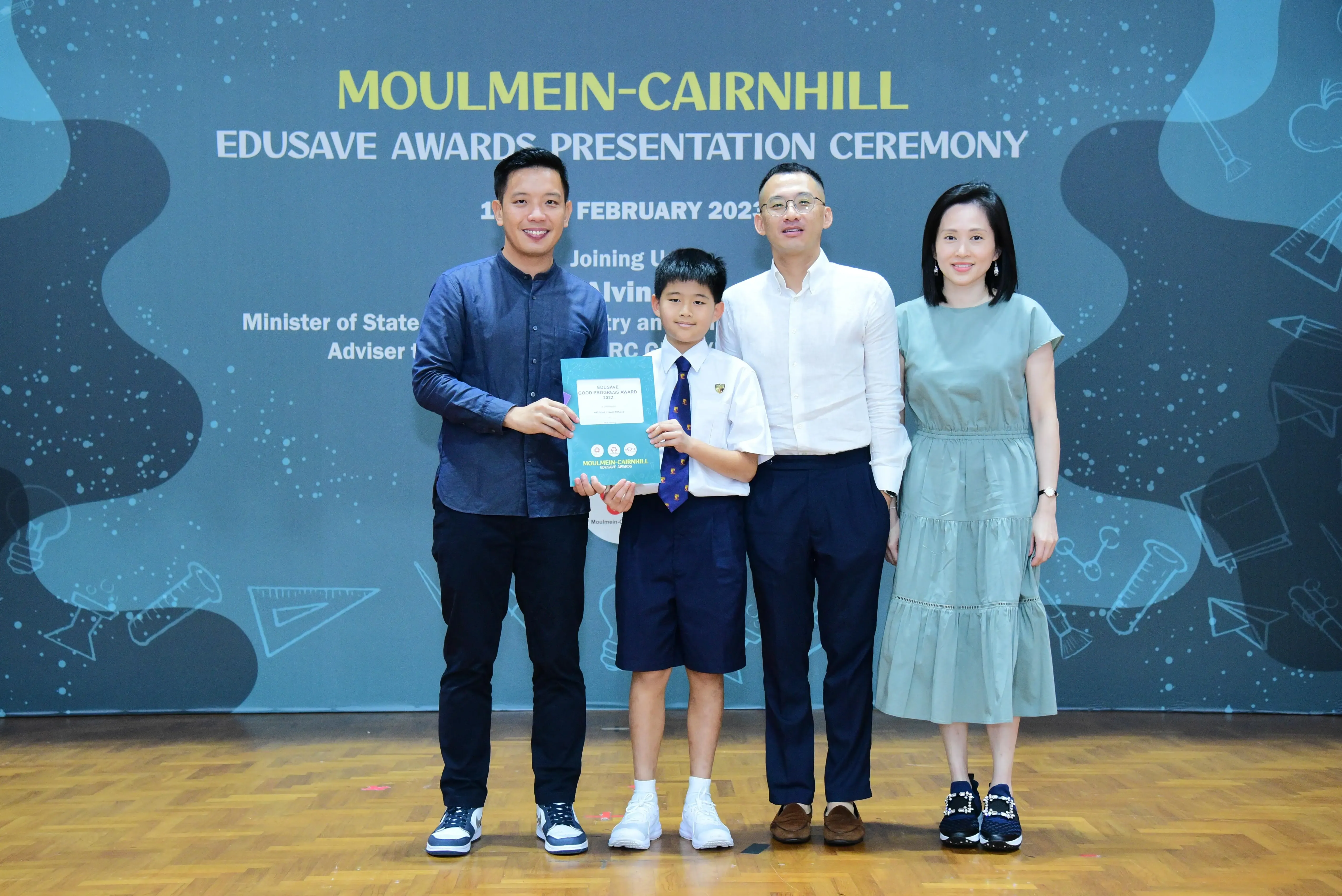 A young student in school uniform holds an award certificate with three adults at the Moulmein-Cairnhill Edusave Awards Ceremony.