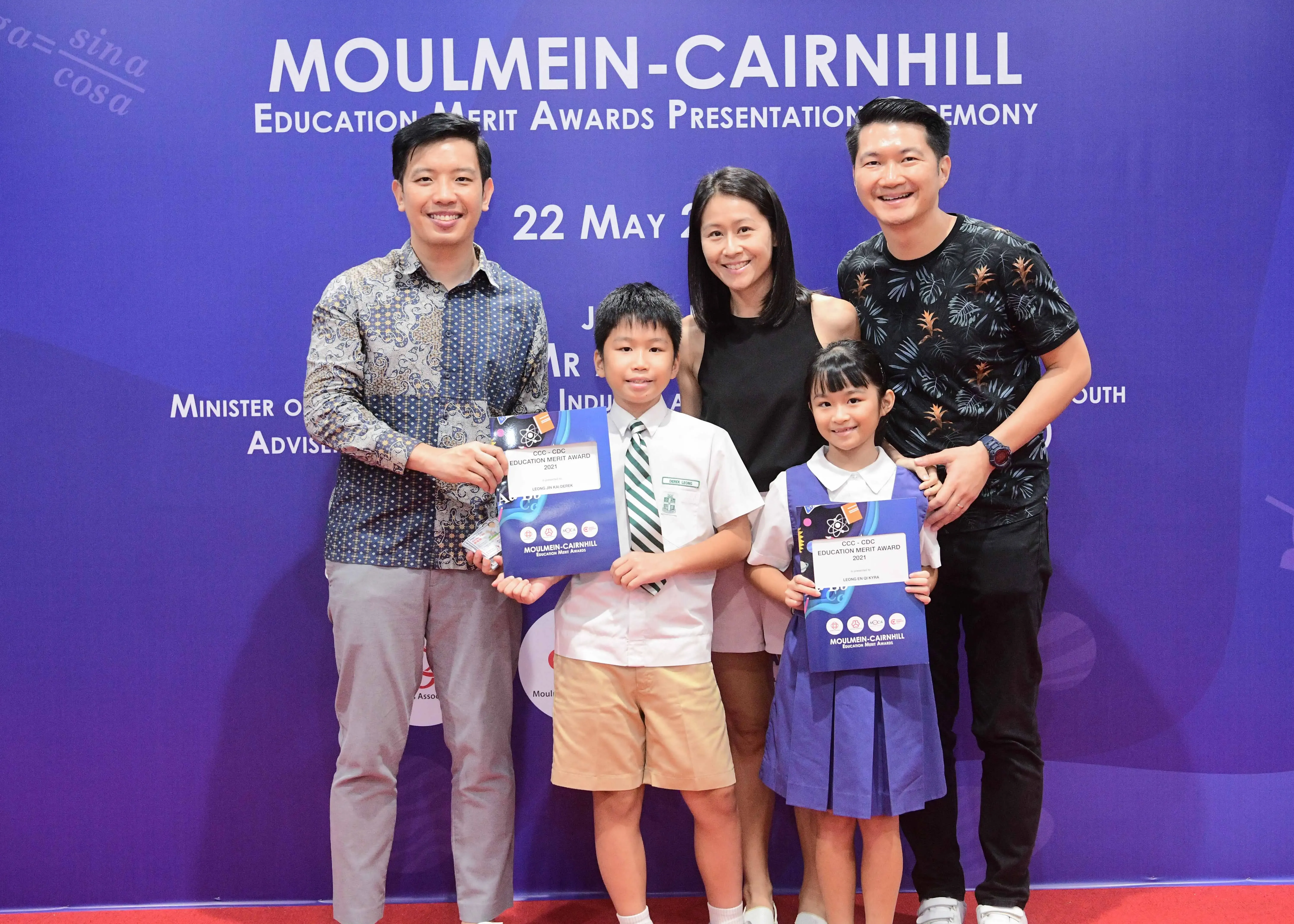Five people pose at the Moulmein-Cairnhill Education Merit Awards ceremony, two children holding award certificates.