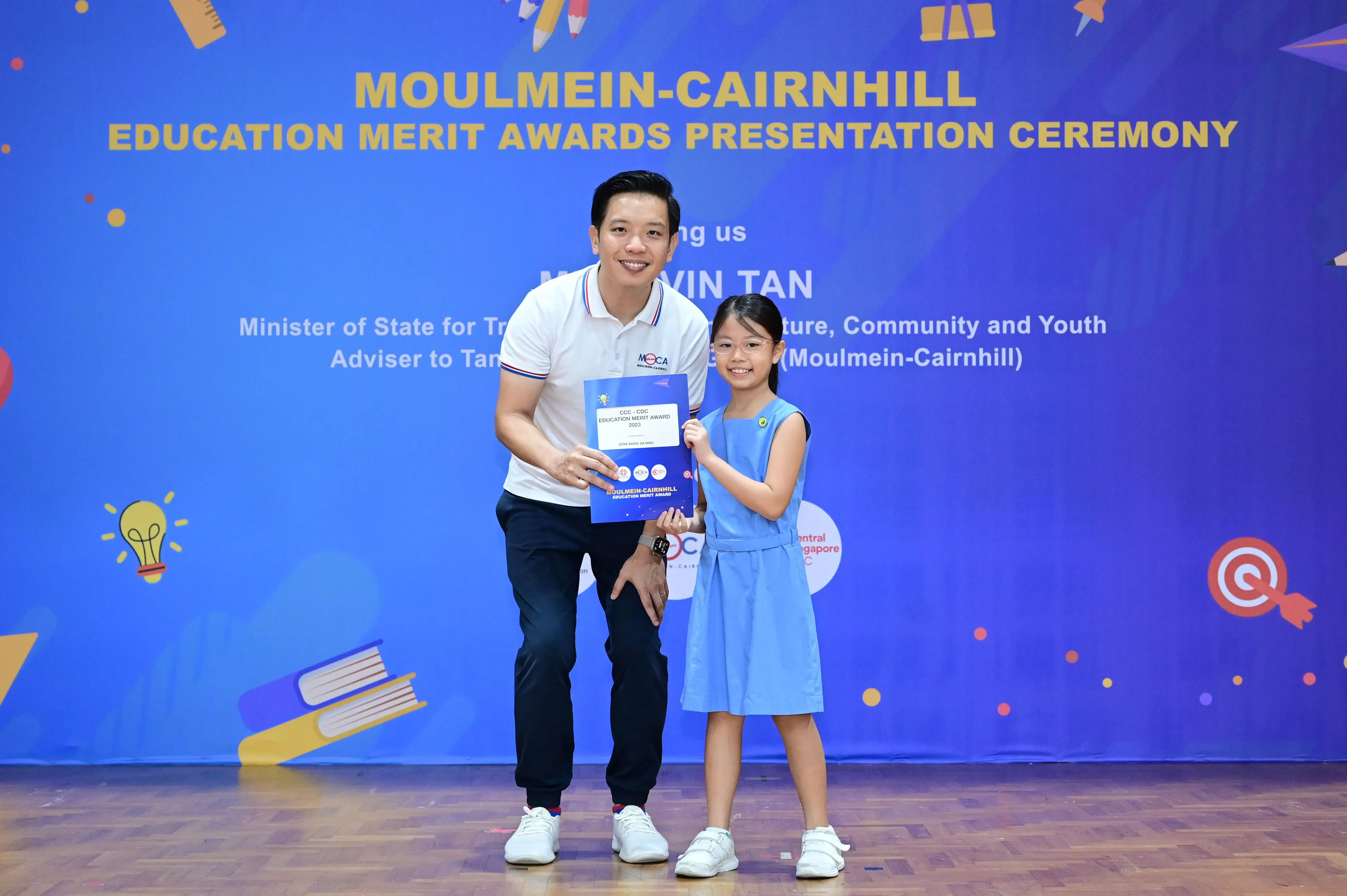 A man and a young girl hold a merit award certificate in front of a Moulmein-Cairnhill Education Merit Awards banner.
