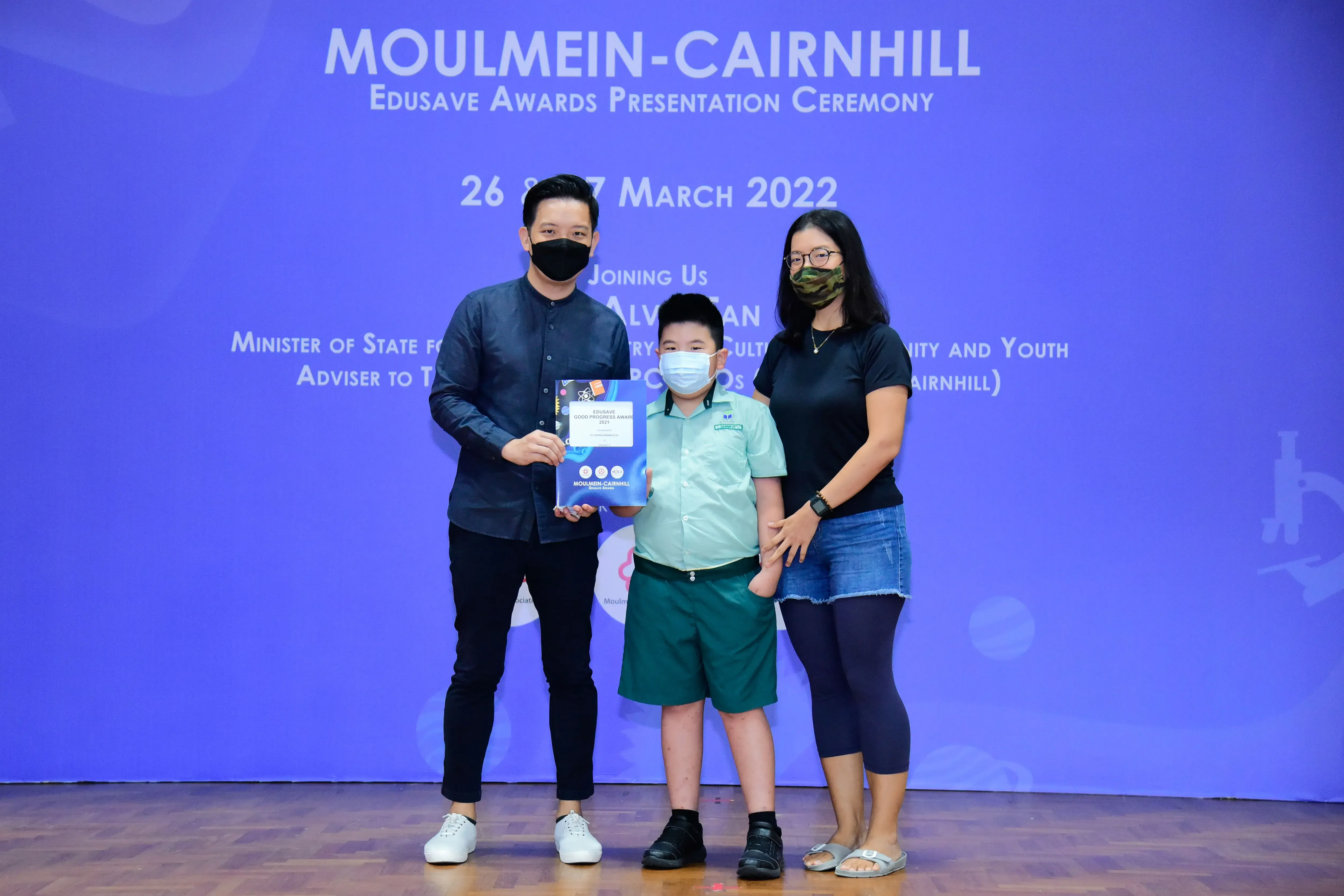 Two masked adults and a masked child in school uniform pose holding an award at the Moulmein-Cairnhill Edusave ceremony.