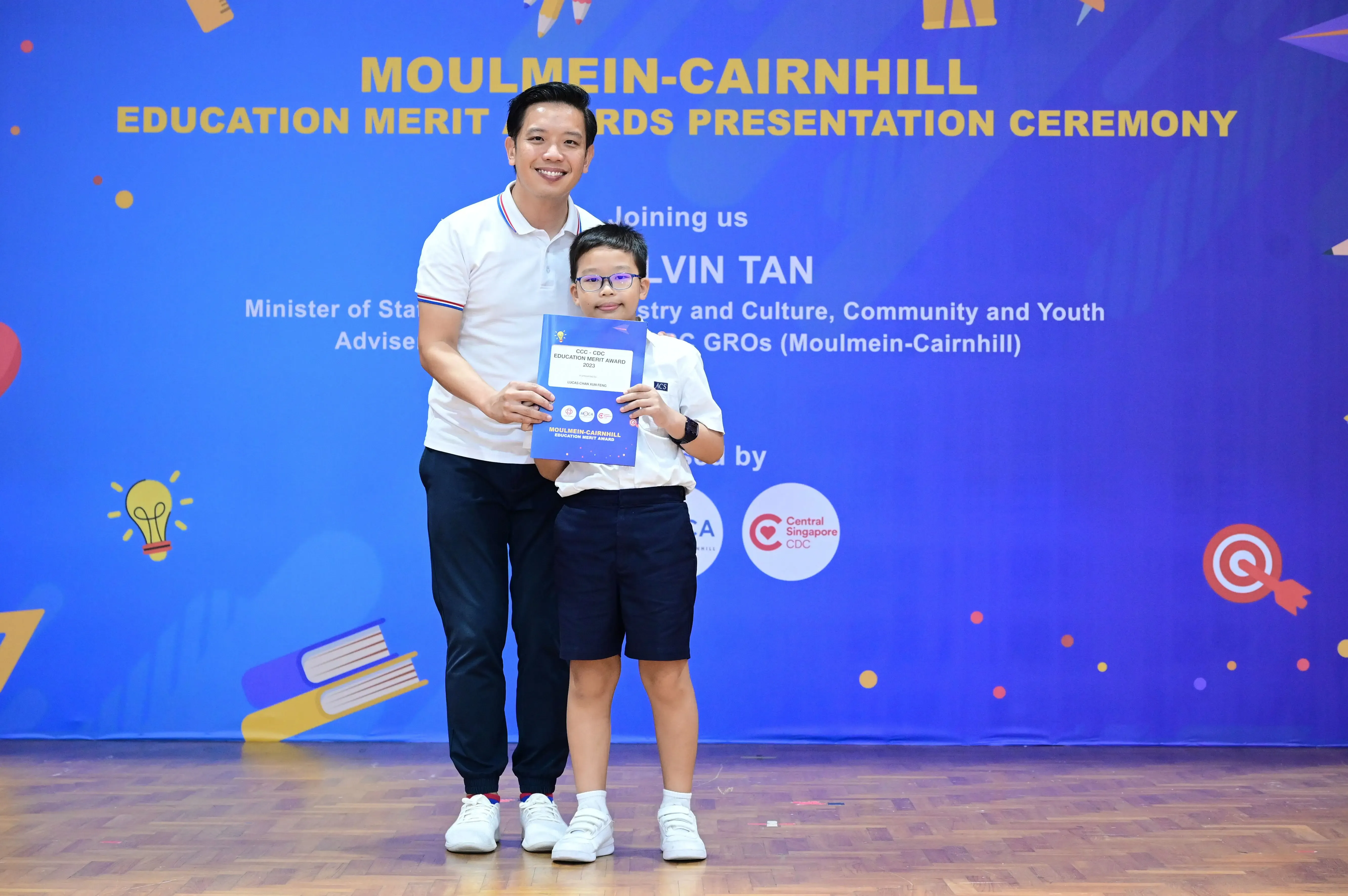 A man and a young boy hold a merit award certificate at the Moulmein-Cairnhill Education Merit Awards Presentation Ceremony.