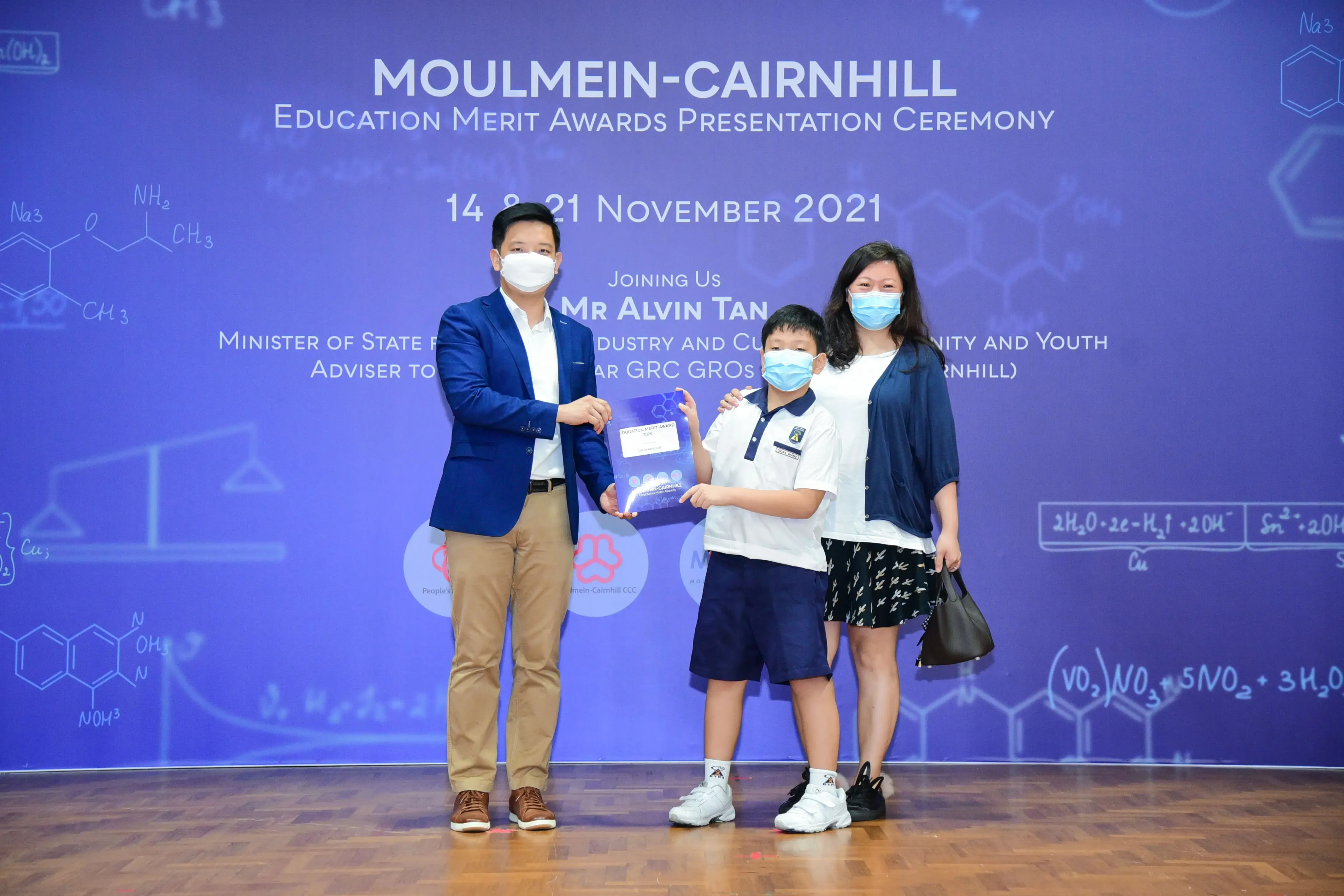 A man in a blue blazer presents an award to a masked boy and woman at the Moulmein-Cairnhill Education Merit Awards 2021.