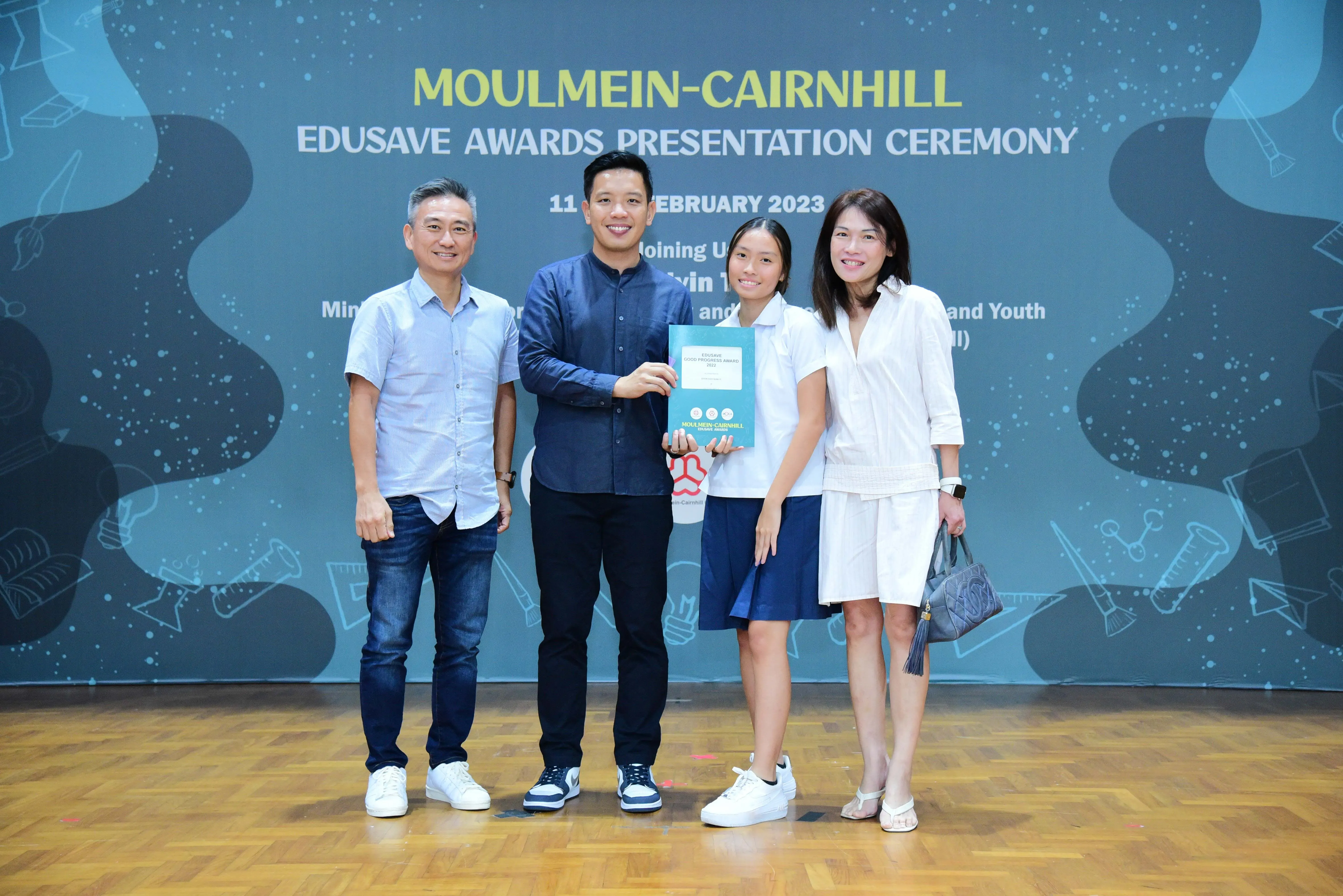 Four people pose at the Moulmein-Cairnhill Edusave Awards Presentation Ceremony, with a student holding a certificate.