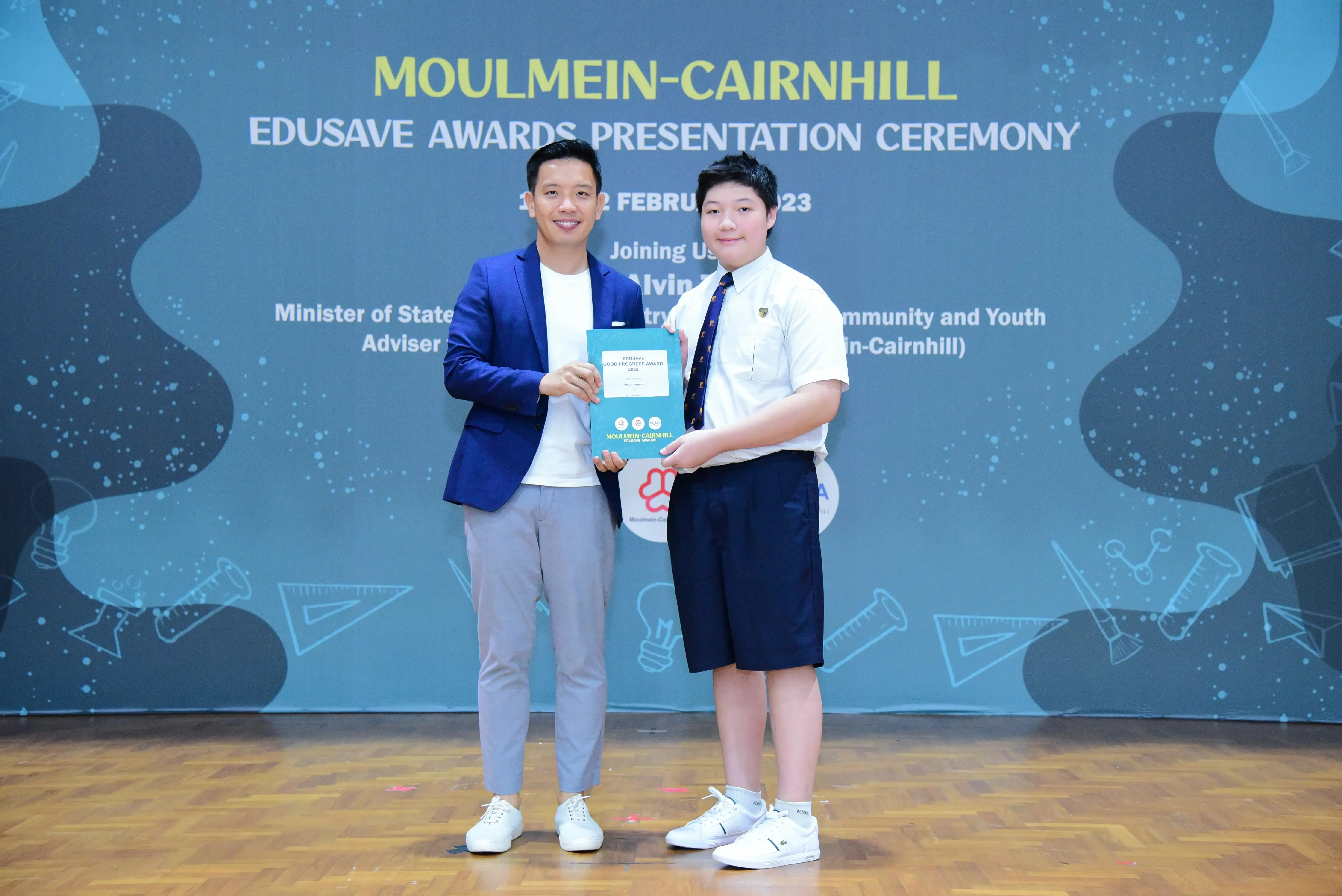 An adult in a blue blazer and a student in school uniform hold an award at the Moulmein-Cairnhill Edusave Awards Ceremony.