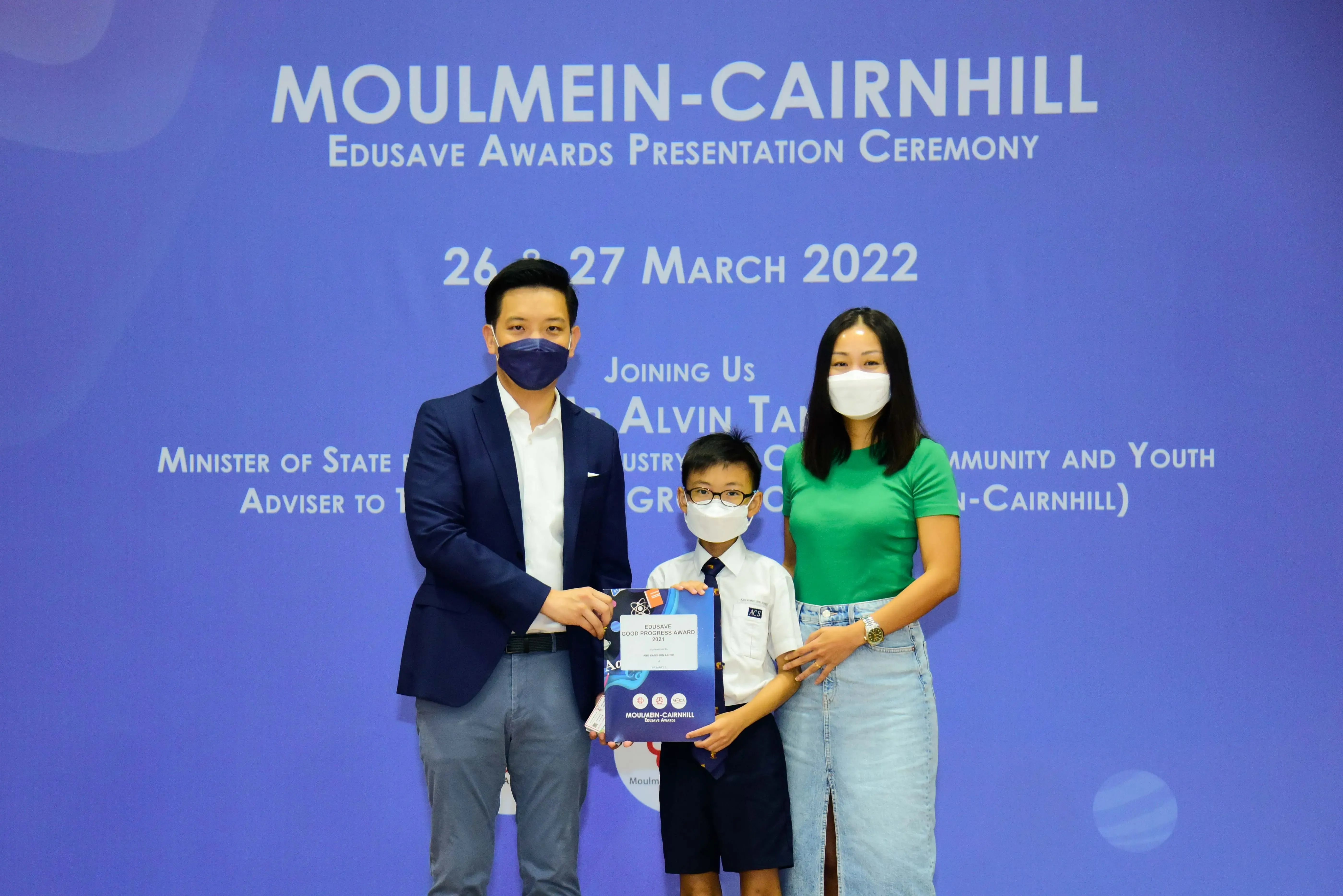 Two masked adults and a masked child hold an award at the Moulmein-Cairnhill Edusave Awards Ceremony, March 2022.