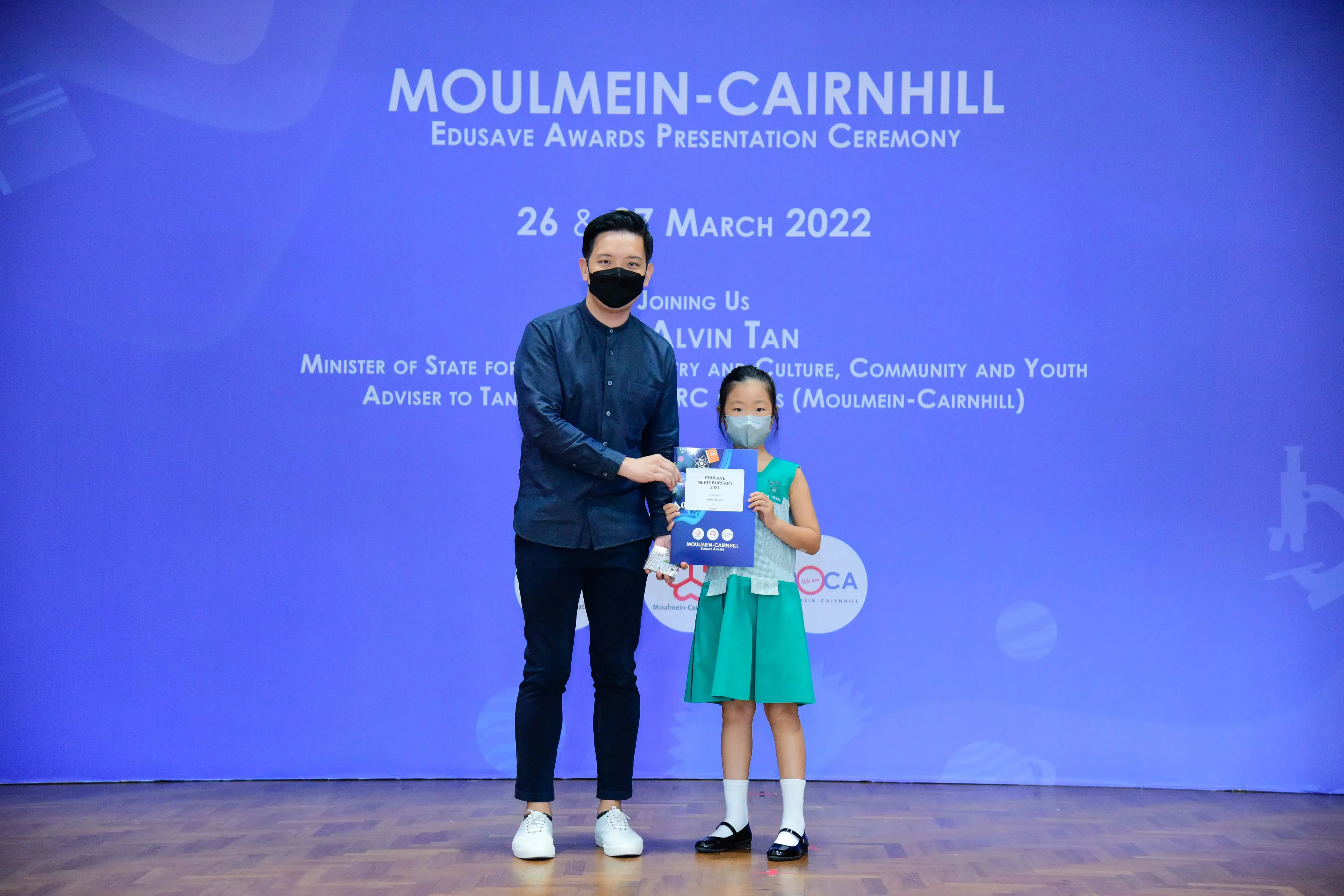 A masked man presents an award to a young girl in a green dress at the Moulmein-Cairnhill Edusave Awards Ceremony 2022.