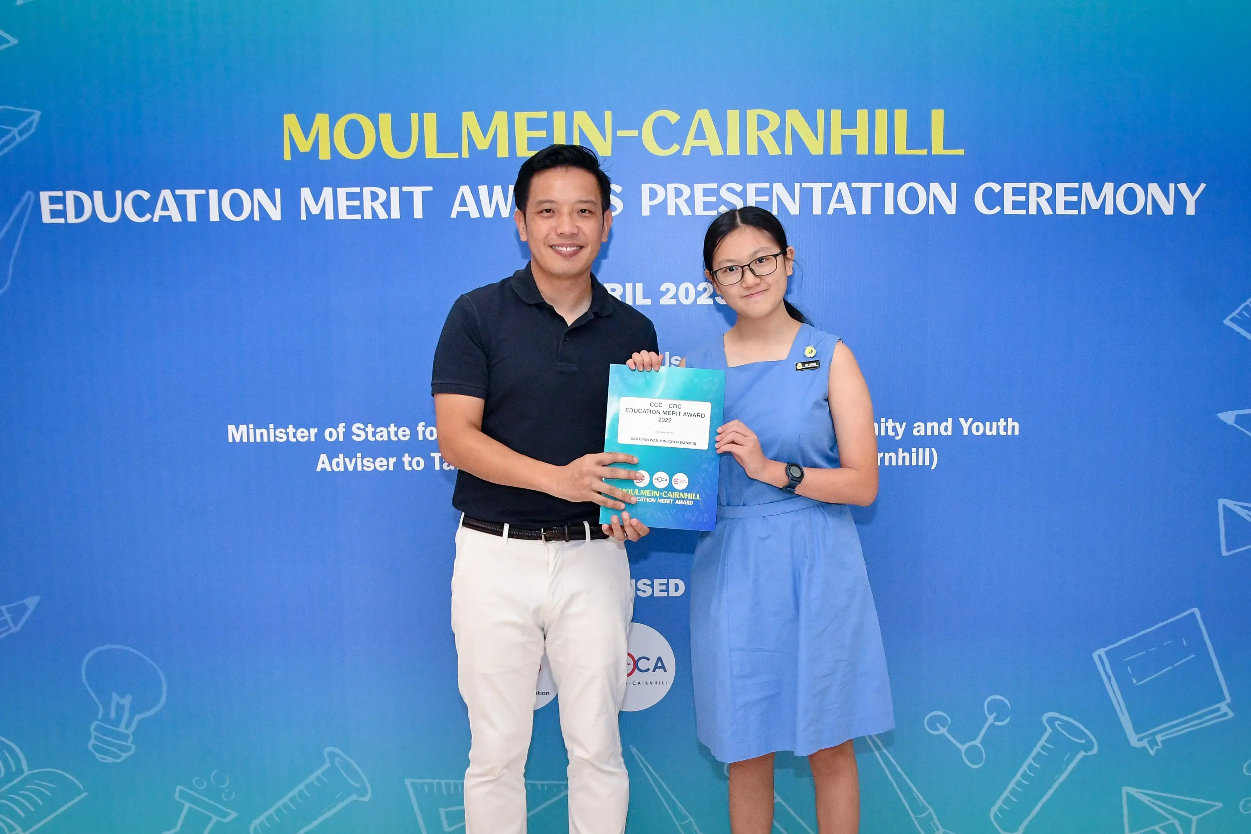 A man and a young girl hold a certificate together in front of a Moulmein-Cairnhill Education Merit Awards banner.