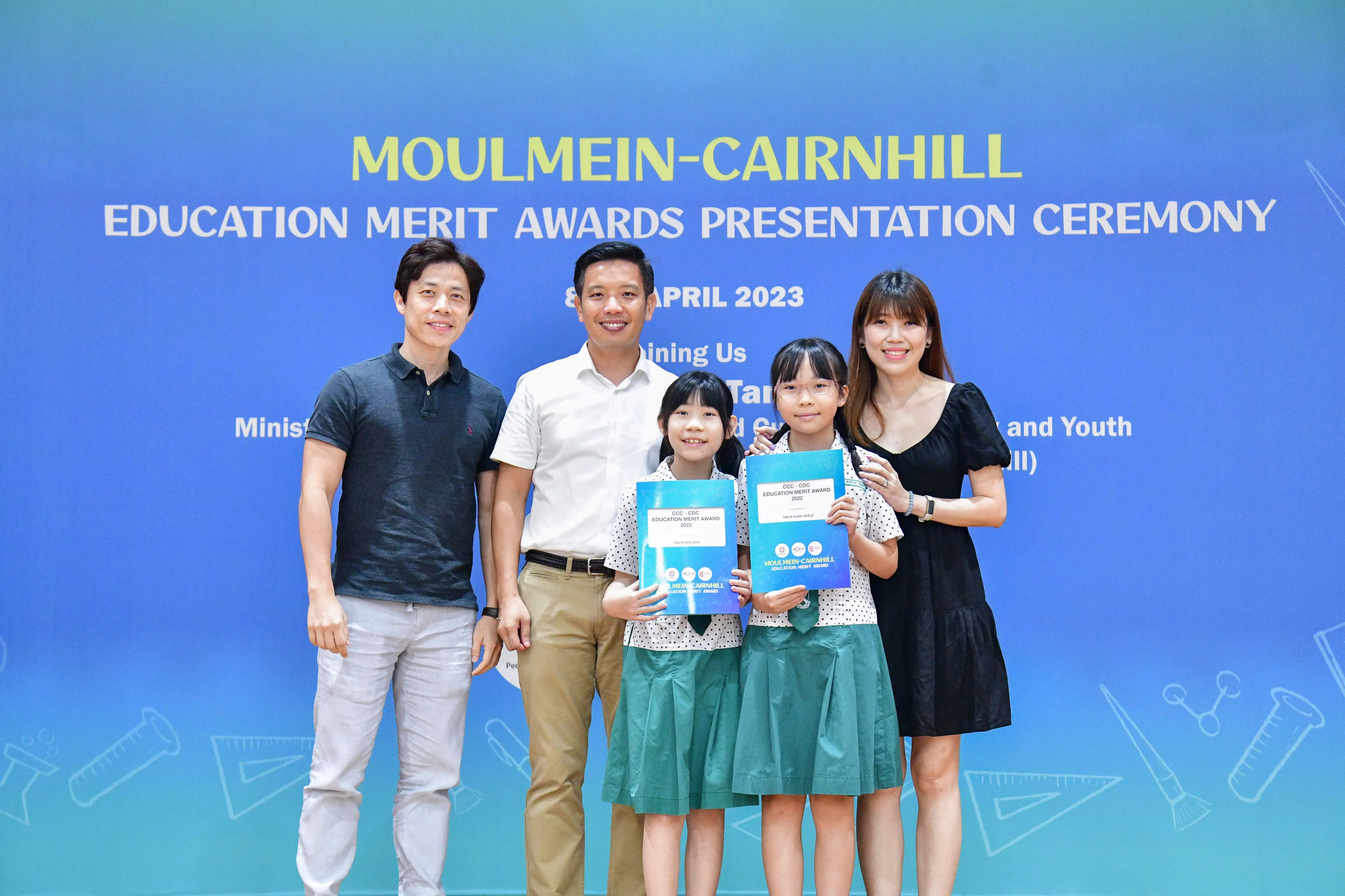Two girls holding award certificates pose with three adults at the Moulmein-Cairnhill Education Merit Awards Ceremony, April 2023.