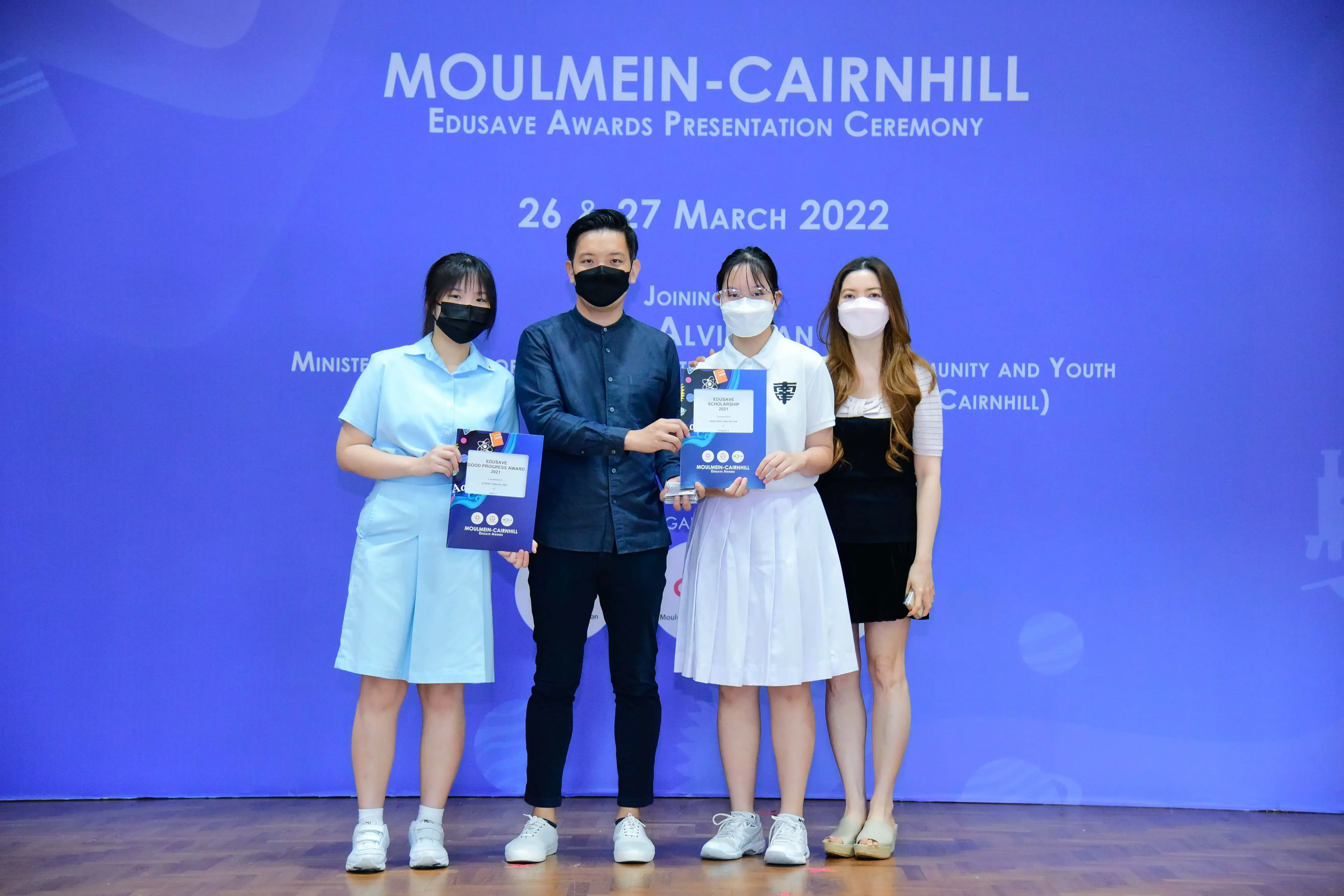 Four masked people pose holding award booklets at the Moulmein-Cairnhill Edusave Awards Ceremony, March 2022.