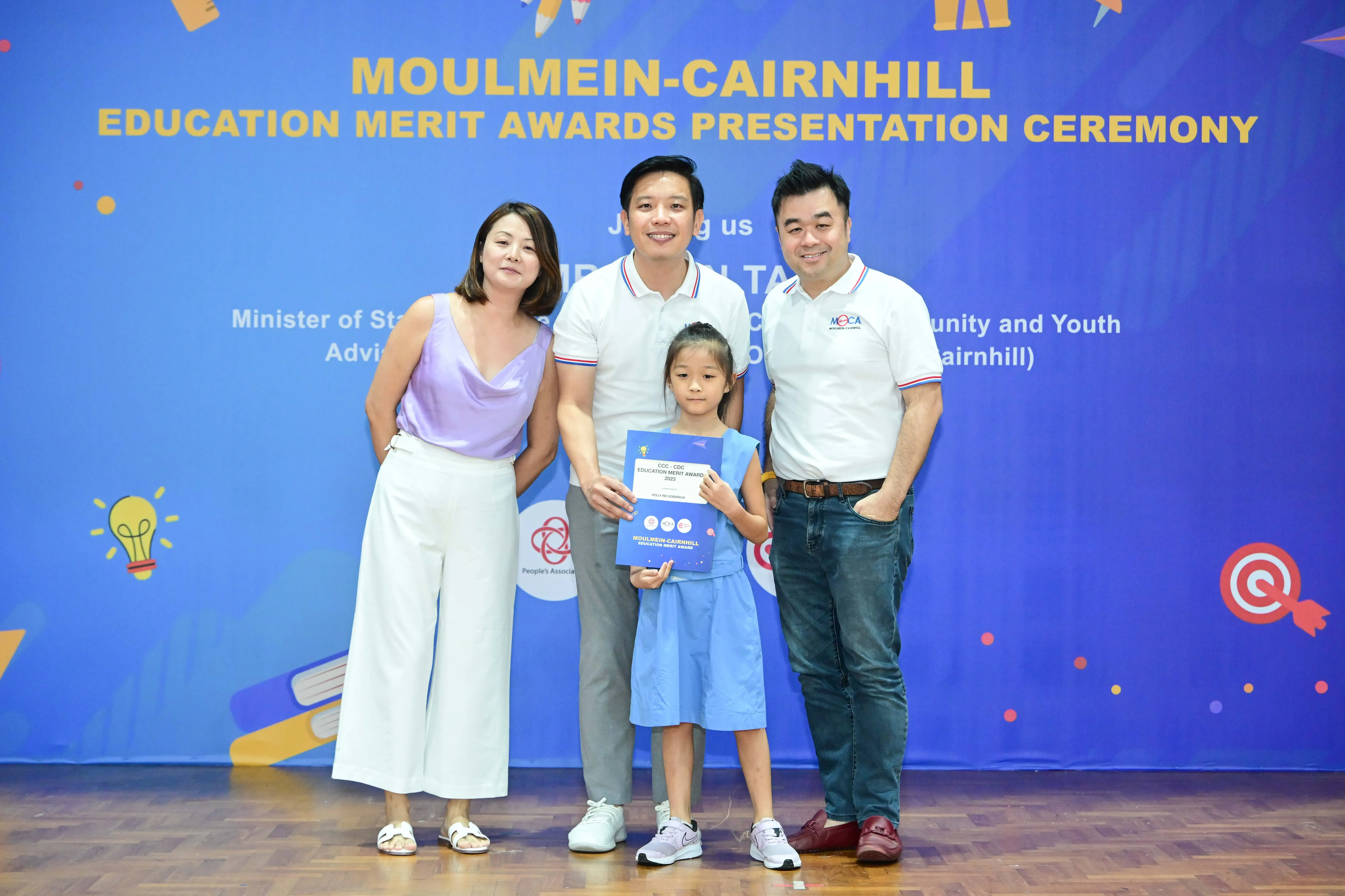 Three adults and a young girl holding a certificate pose before a Moulmein-Cairnhill Education Merit Awards banner.