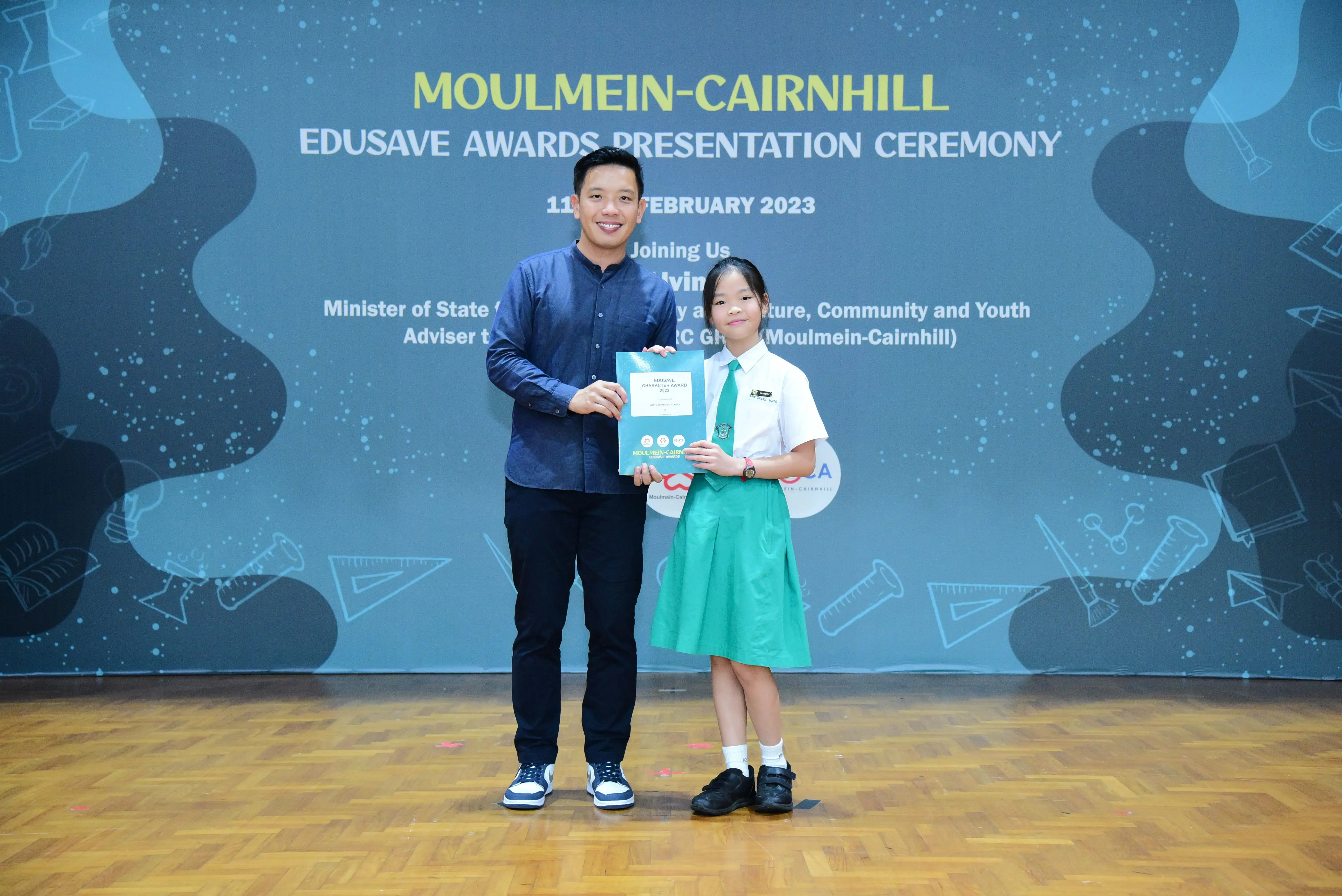 A man and a young girl in school uniform hold a certificate at the Moulmein-Cairnhill Edusave Awards Ceremony 2023.