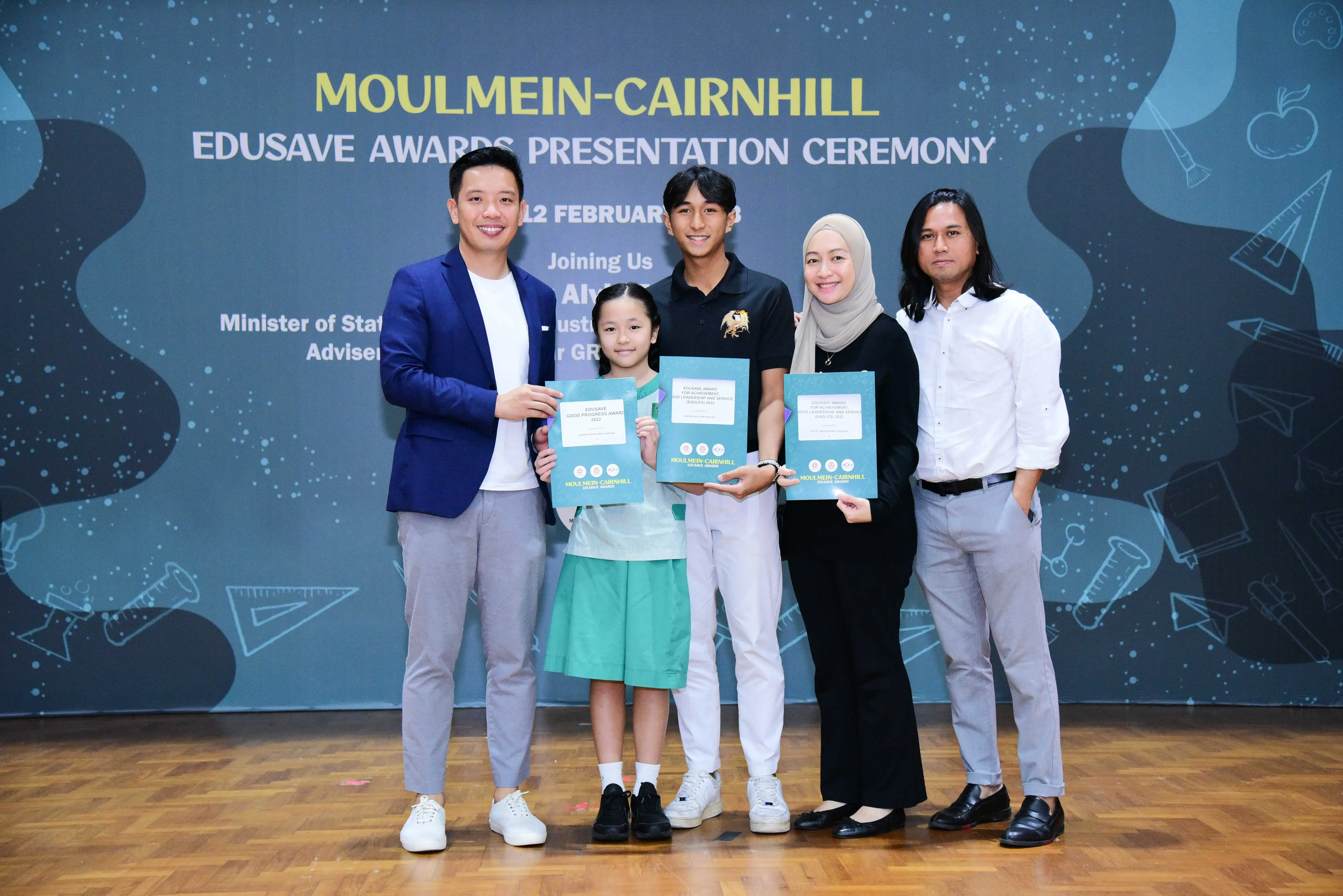 Five people pose holding teal award folders at the Moulmein-Cairnhill Edusave Awards Presentation Ceremony backdrop.