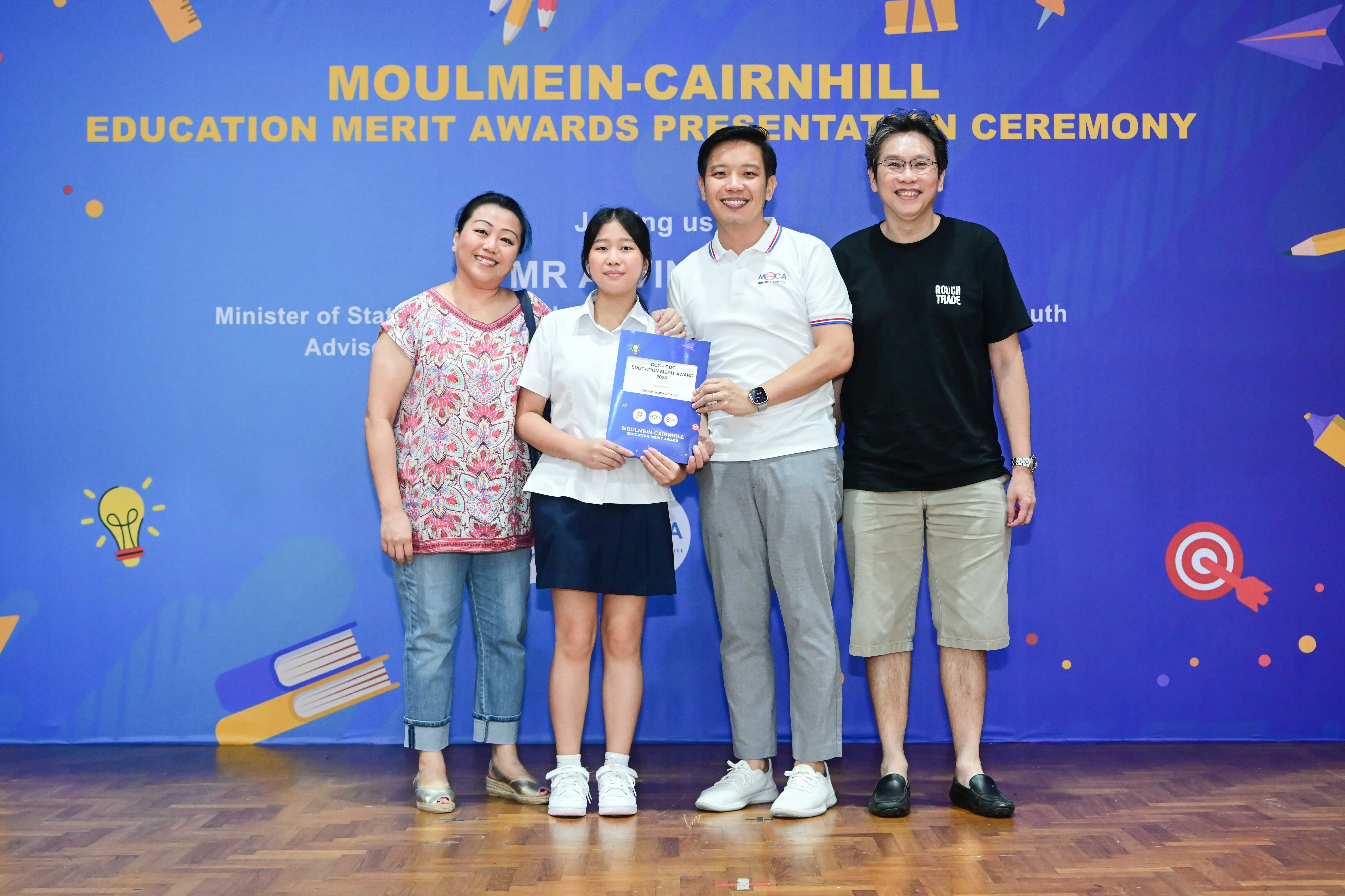 Four people pose smiling at the Moulmein-Cairnhill Education Merit Awards Presentation Ceremony, student holding an award folder.