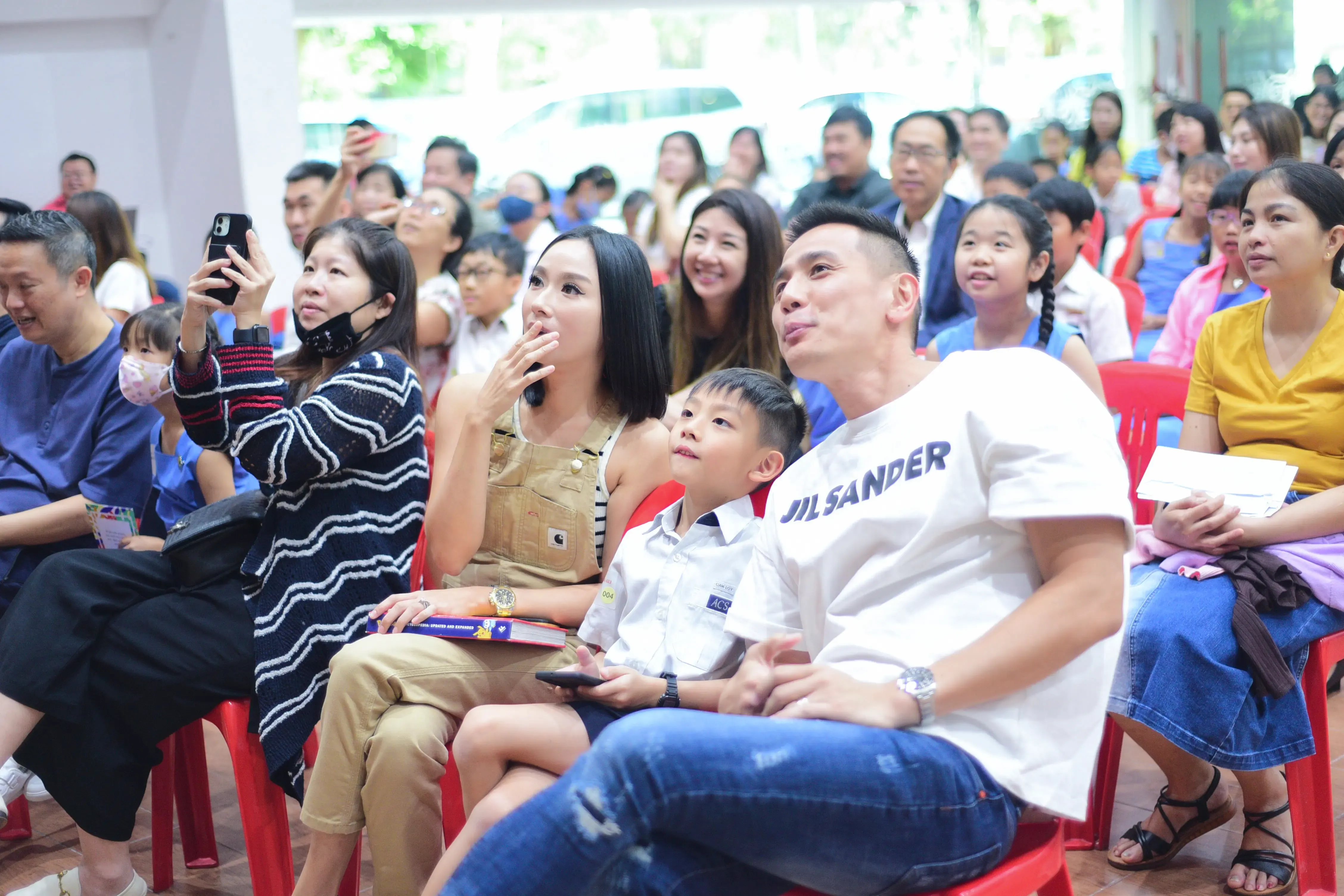 A crowd of adults and children sit on red chairs at an indoor event, looking upward attentively.