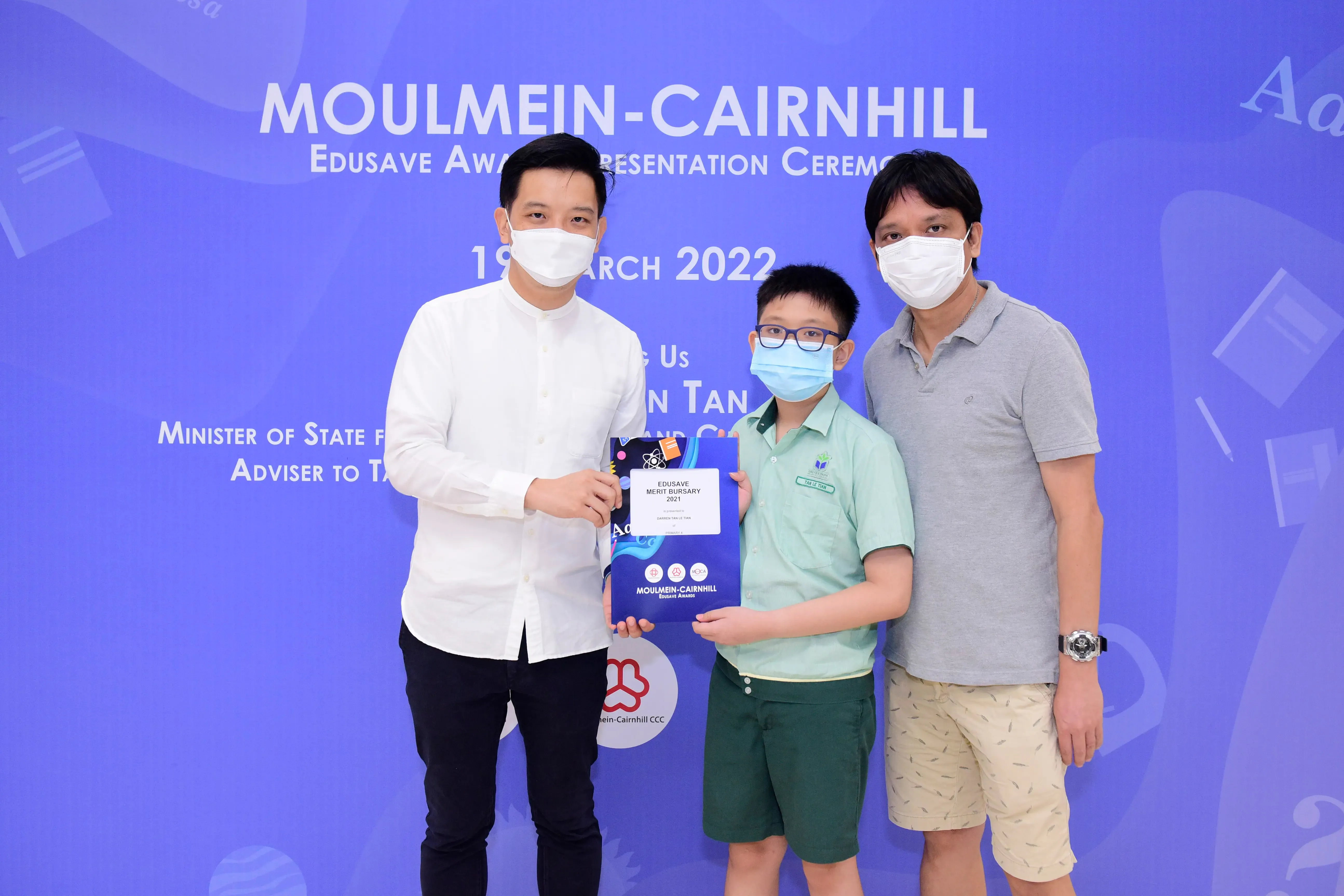 Two masked men and a masked boy hold an Edusave Merit Bursary certificate at the Moulmein-Cairnhill ceremony, March 2022.