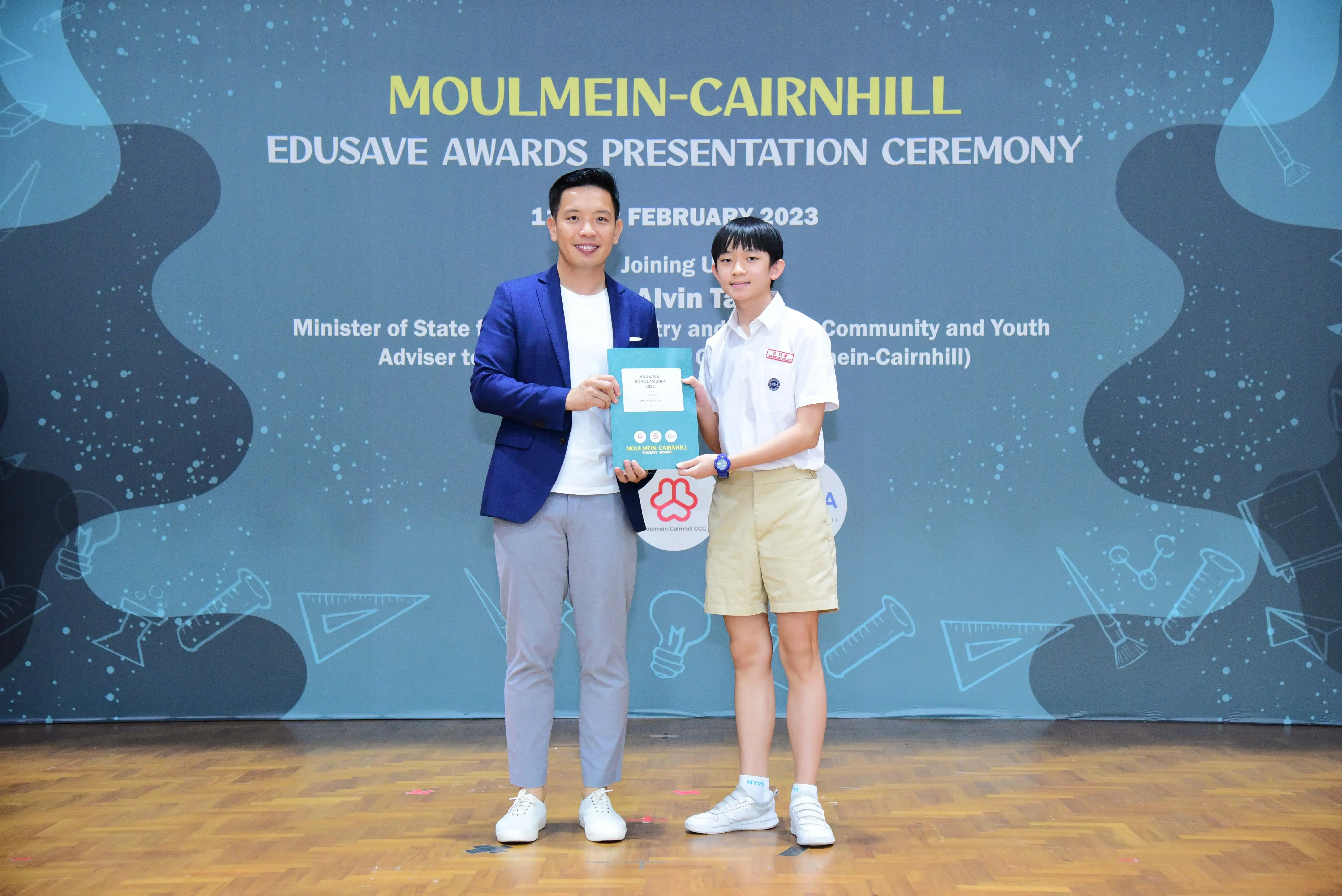 An adult in a blue blazer and a young student in school uniform hold a certificate at the Moulmein-Cairnhill Edusave Awards 2023.