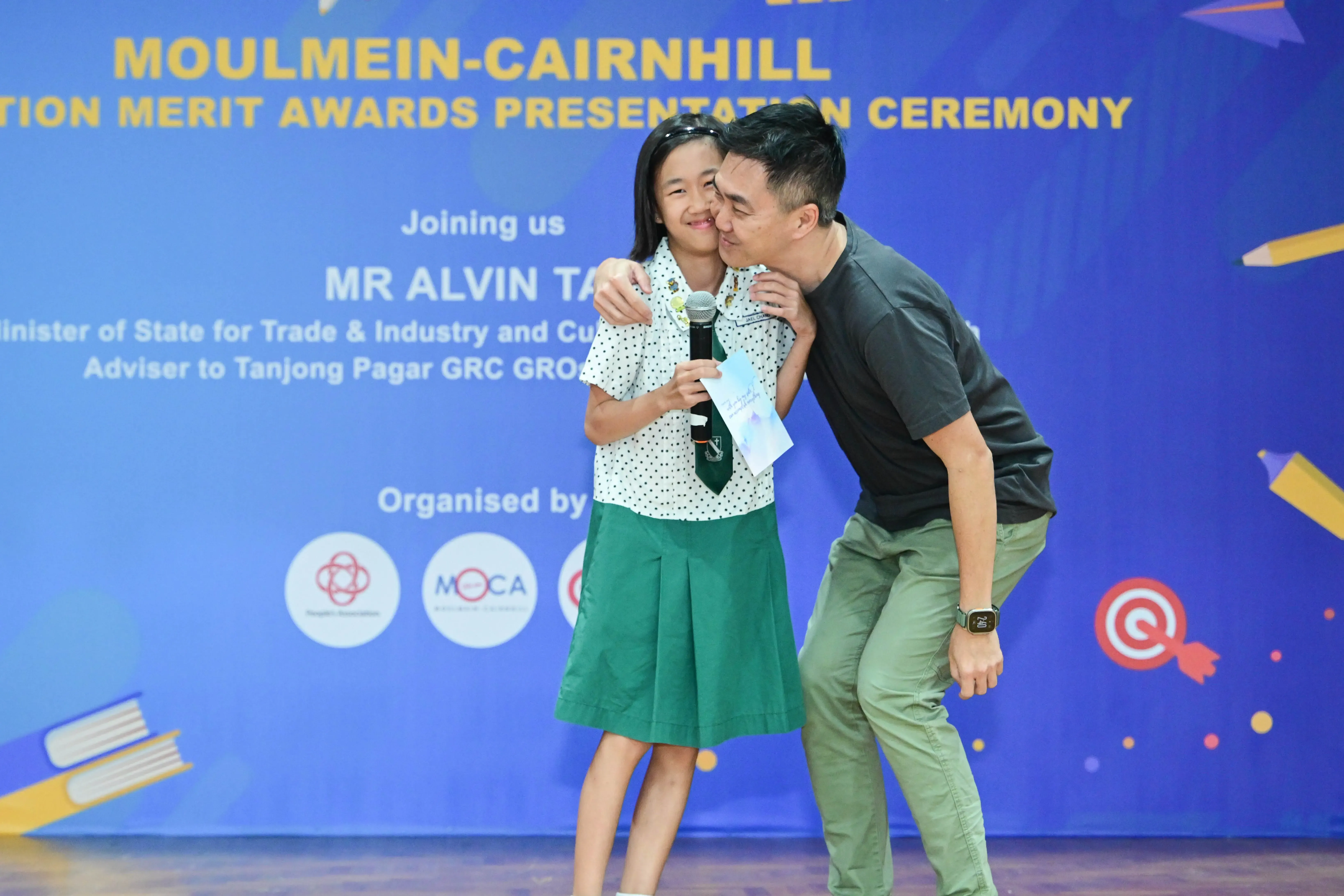 A man kisses a smiling girl holding a microphone at the Moulmein-Cairnhill Merit Awards Presentation Ceremony.