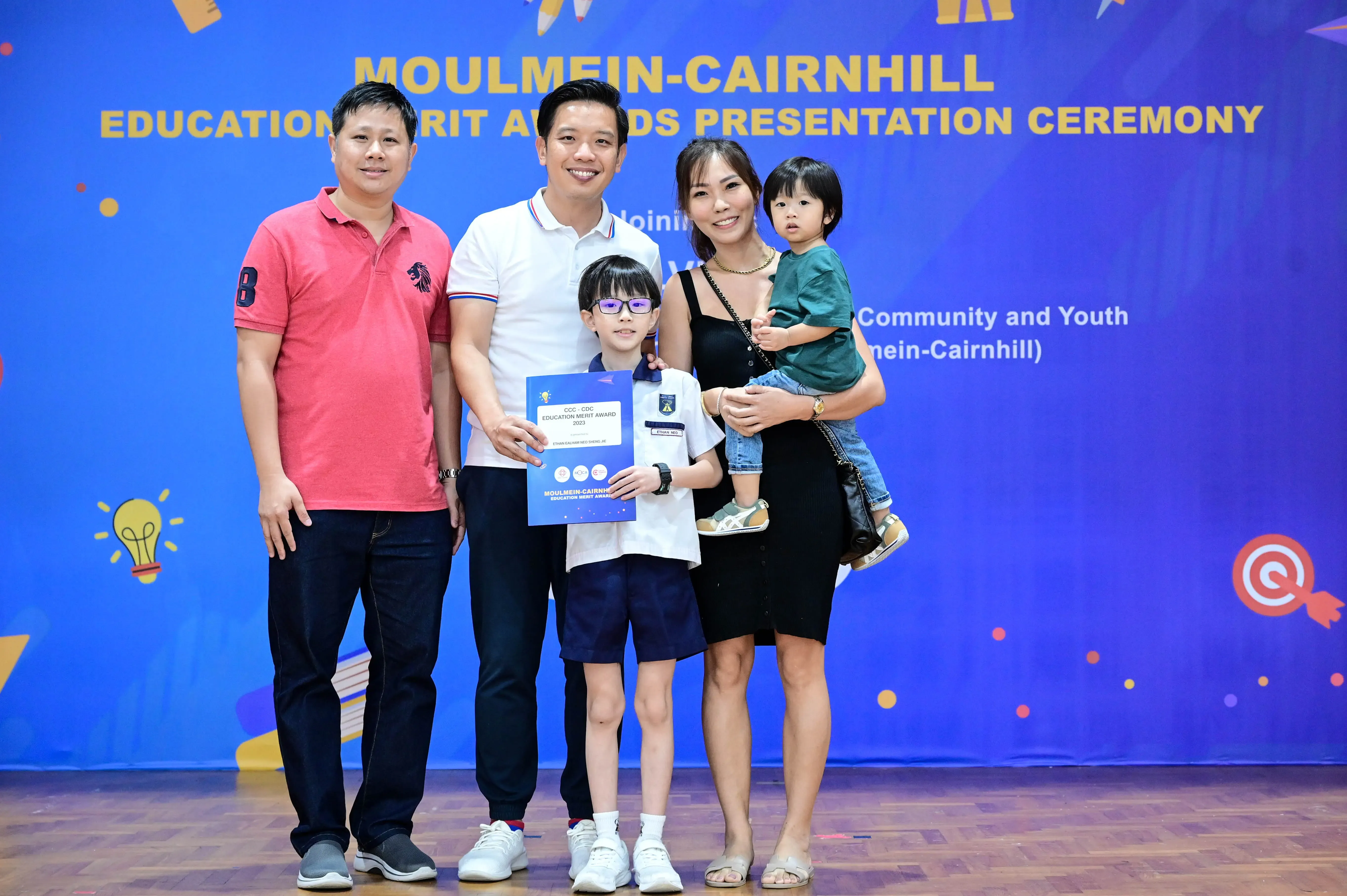 Family posing at Moulmein-Cairnhill Education Merit Awards ceremony, child holding a blue award certificate.