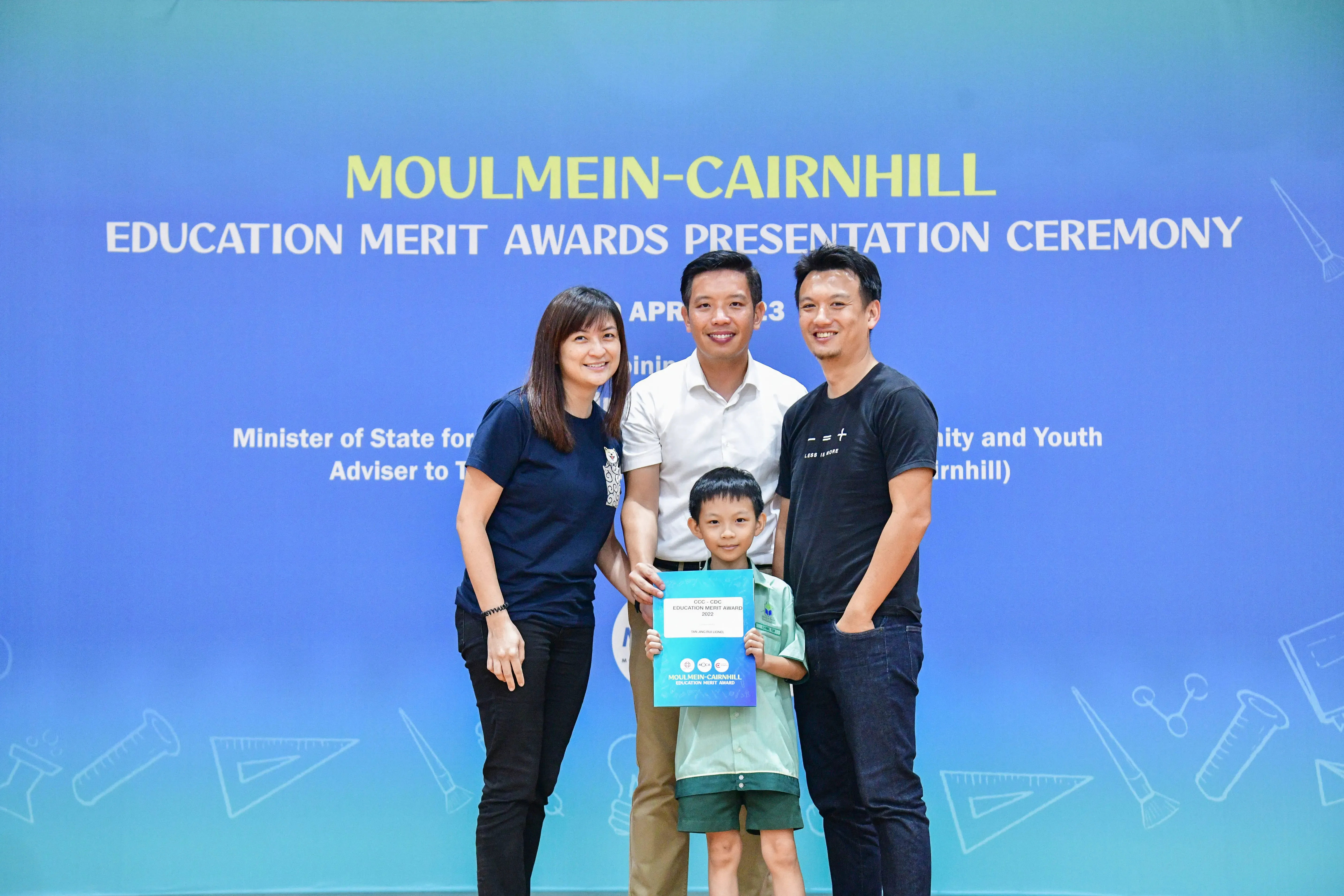 Three adults and a young boy holding an award certificate pose before a Moulmein-Cairnhill Education Merit Awards banner.