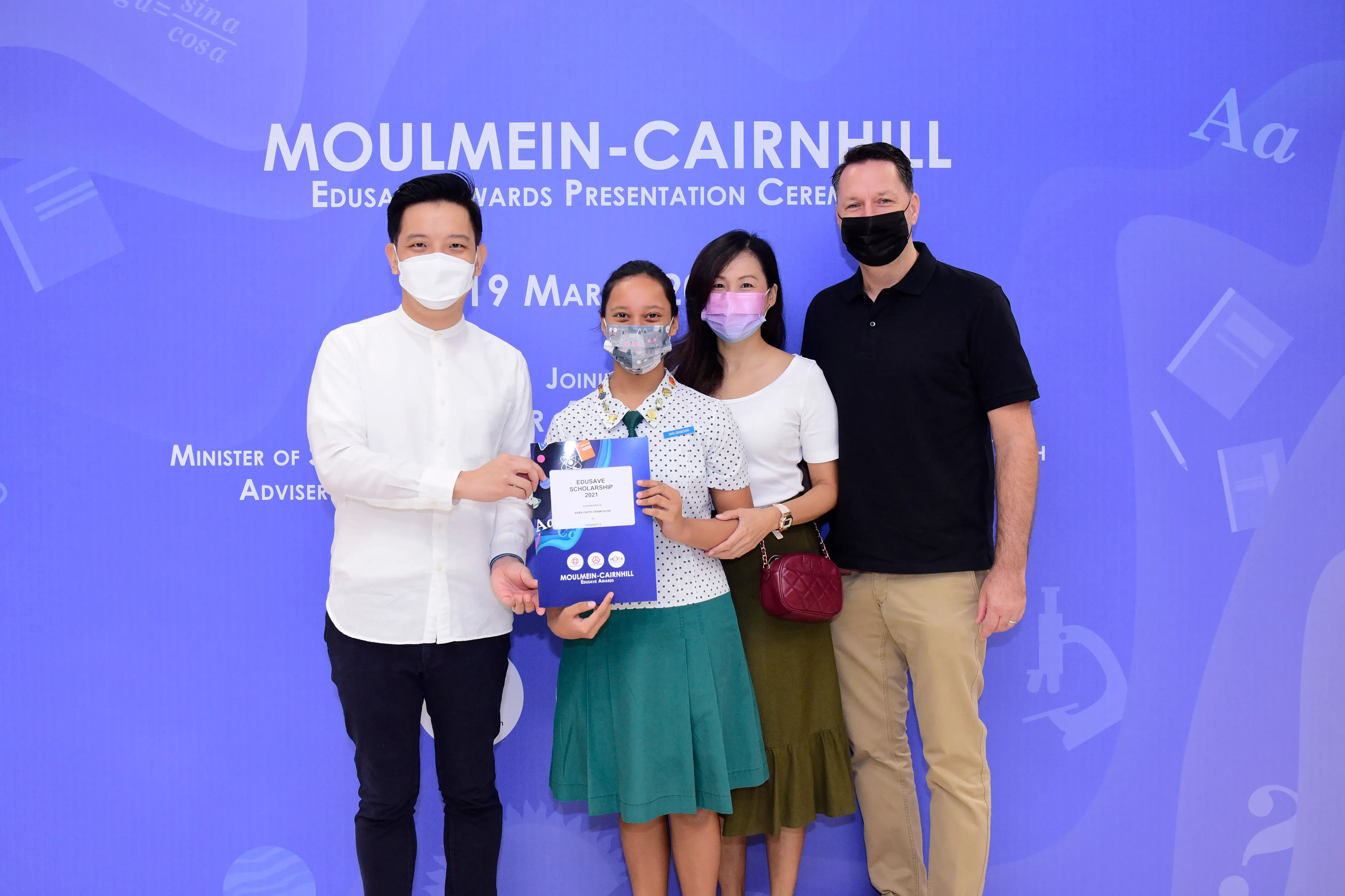 Four masked people pose at the Moulmein-Cairnhill Edusave Awards ceremony, with a student holding a scholarship certificate.