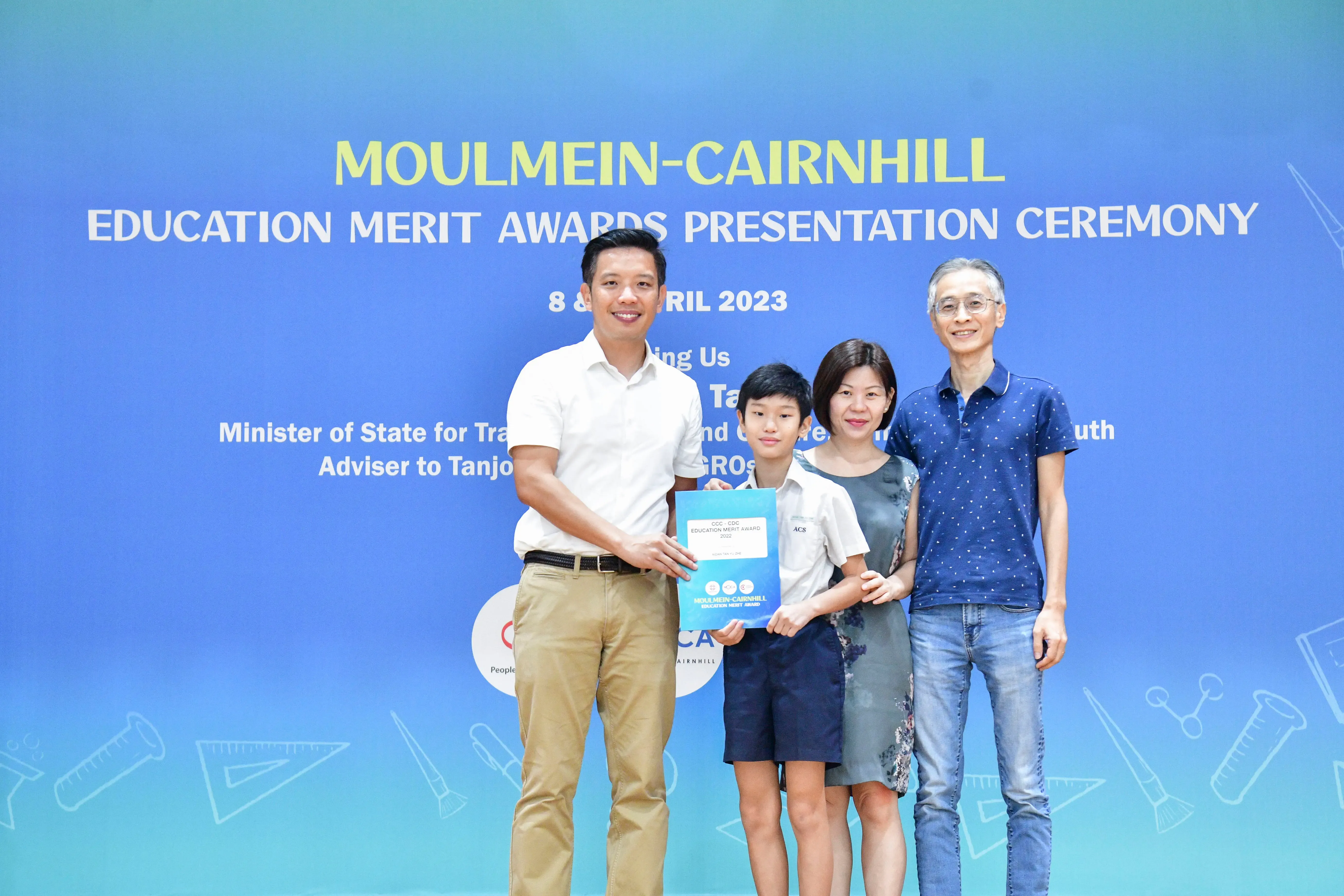 Four people pose at the Moulmein-Cairnhill Education Merit Awards Presentation Ceremony, a boy holding a certificate.