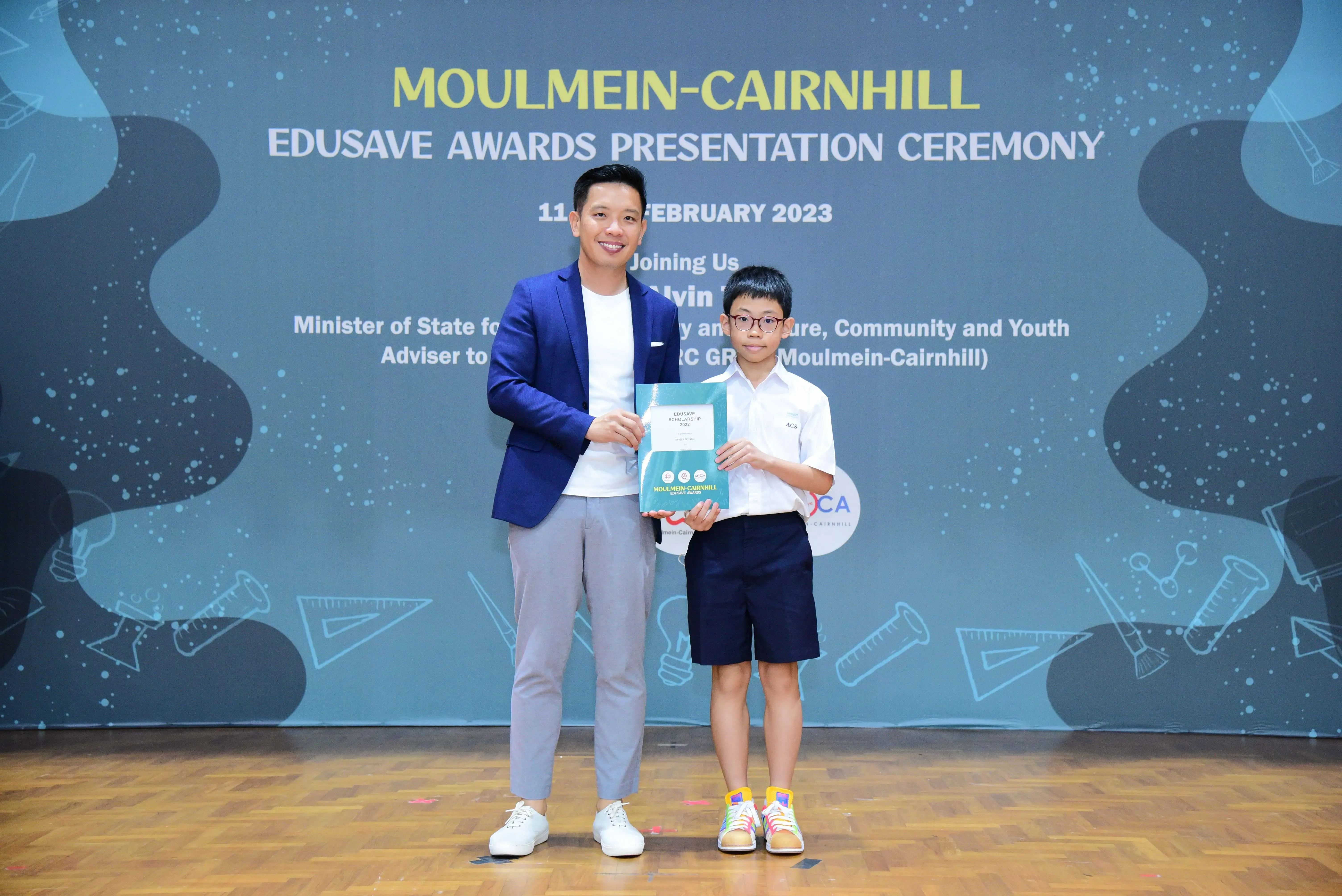 A man in a blue blazer and a young boy holding an award certificate at the Moulmein-Cairnhill Edusave Awards Ceremony 2023.