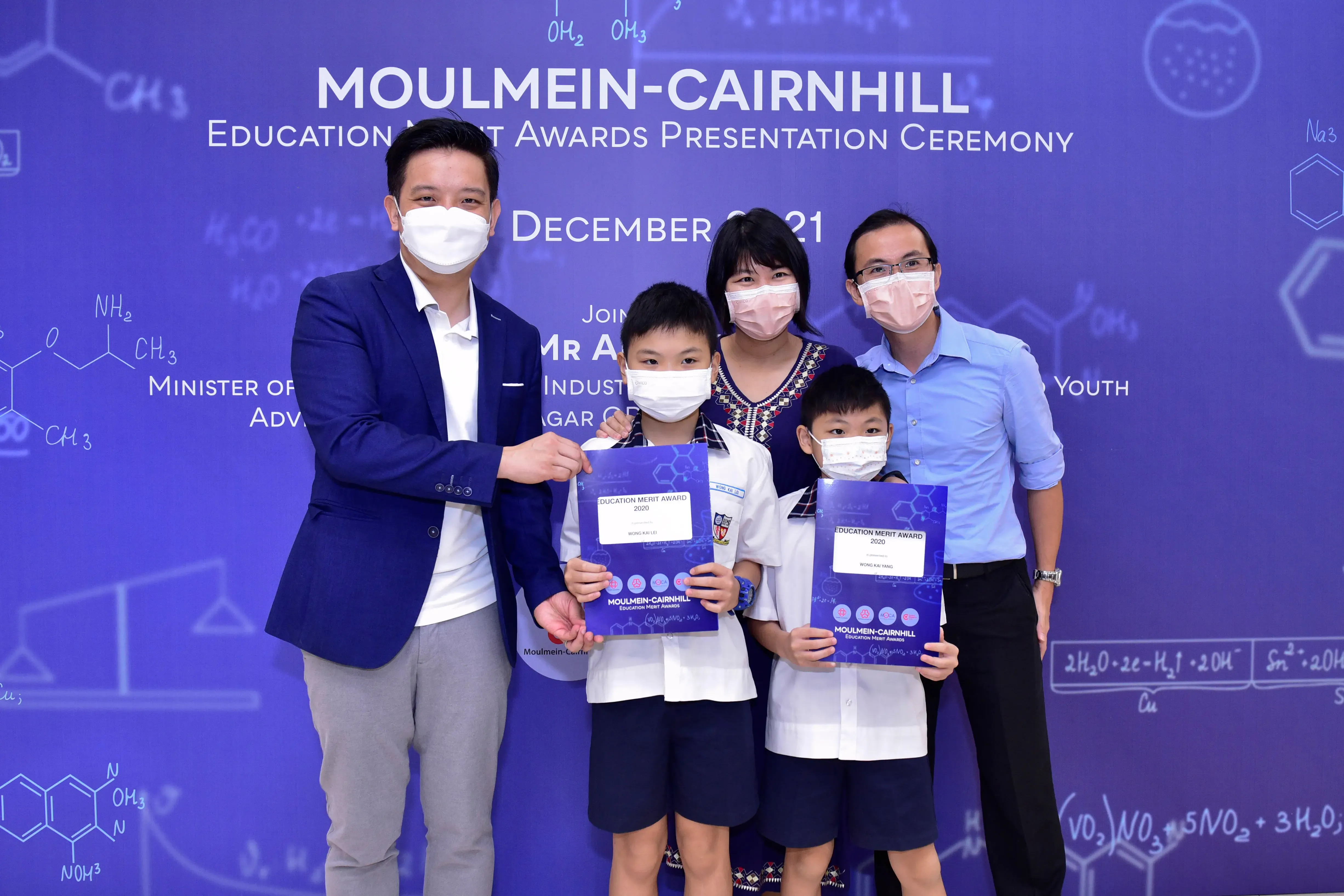 Five masked people pose at Moulmein-Cairnhill Education Merit Awards Ceremony, two children holding award certificates.