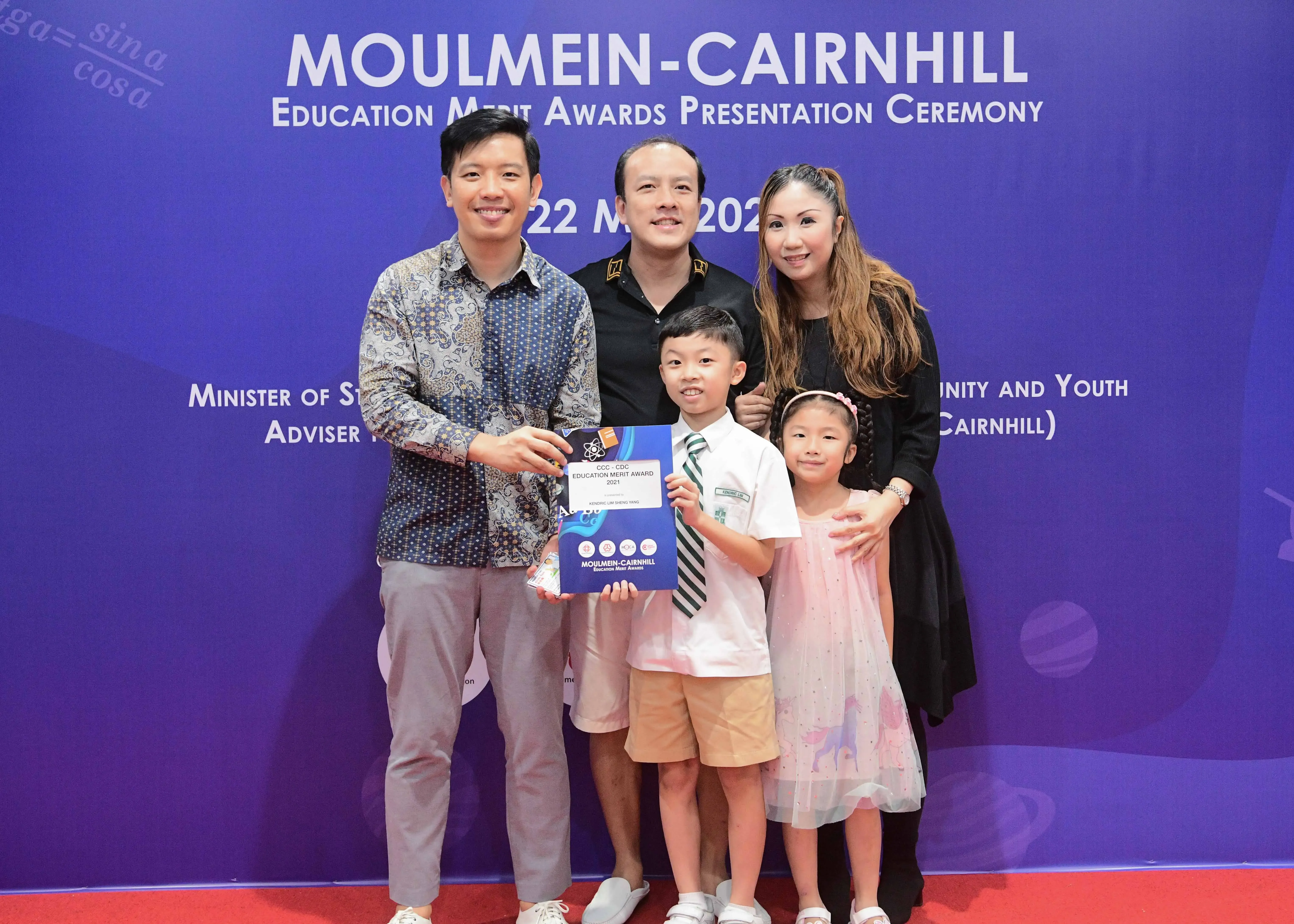 Five people pose before a Moulmein-Cairnhill Education Merit Awards banner; a boy holds an award certificate.