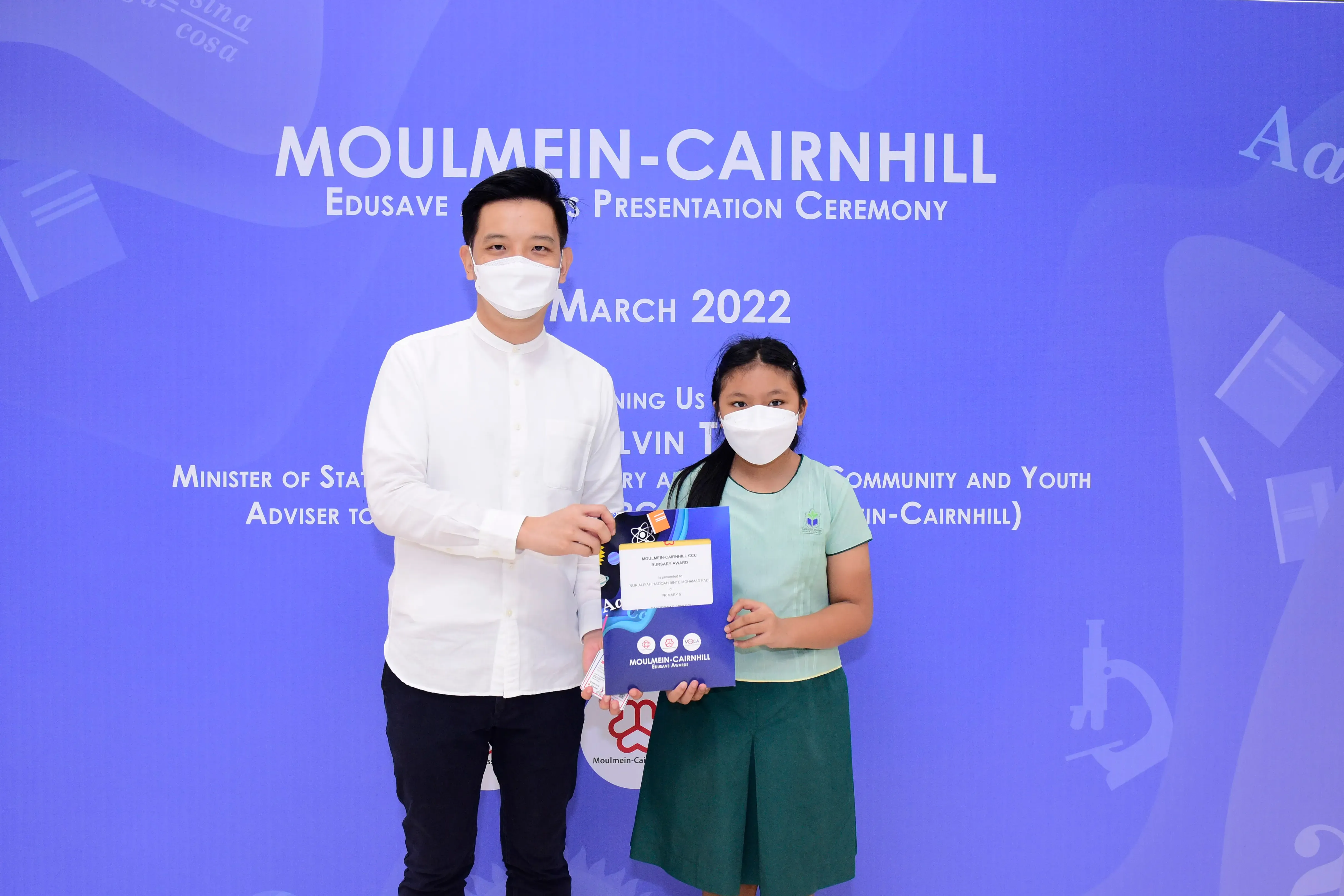 A masked man and a young girl hold an award certificate at the Moulmein-Cairnhill Edusave Presentation Ceremony, March 2022.