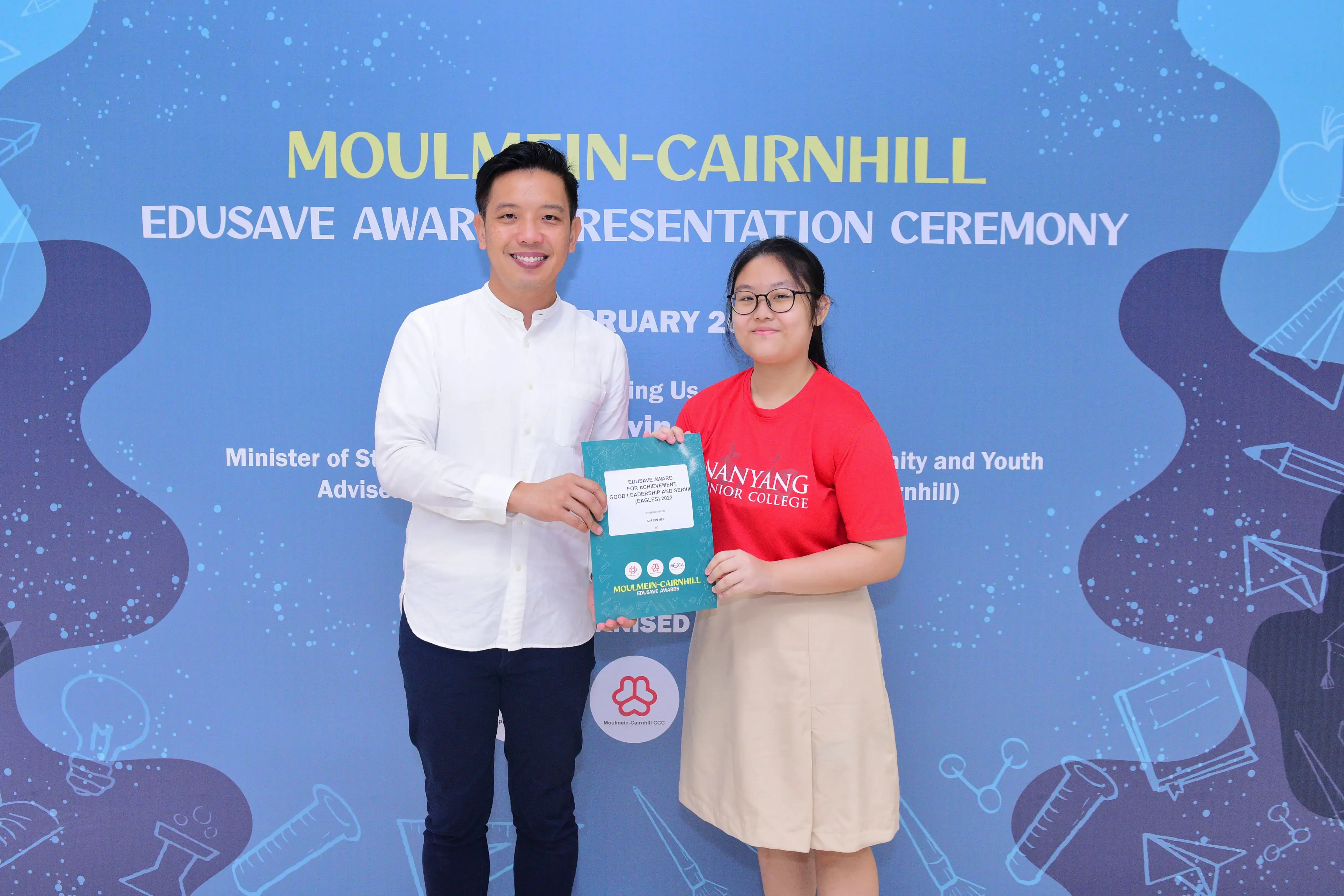 Two people hold a certificate together at the Moulmein-Cairnhill Edusave Award Presentation Ceremony backdrop.