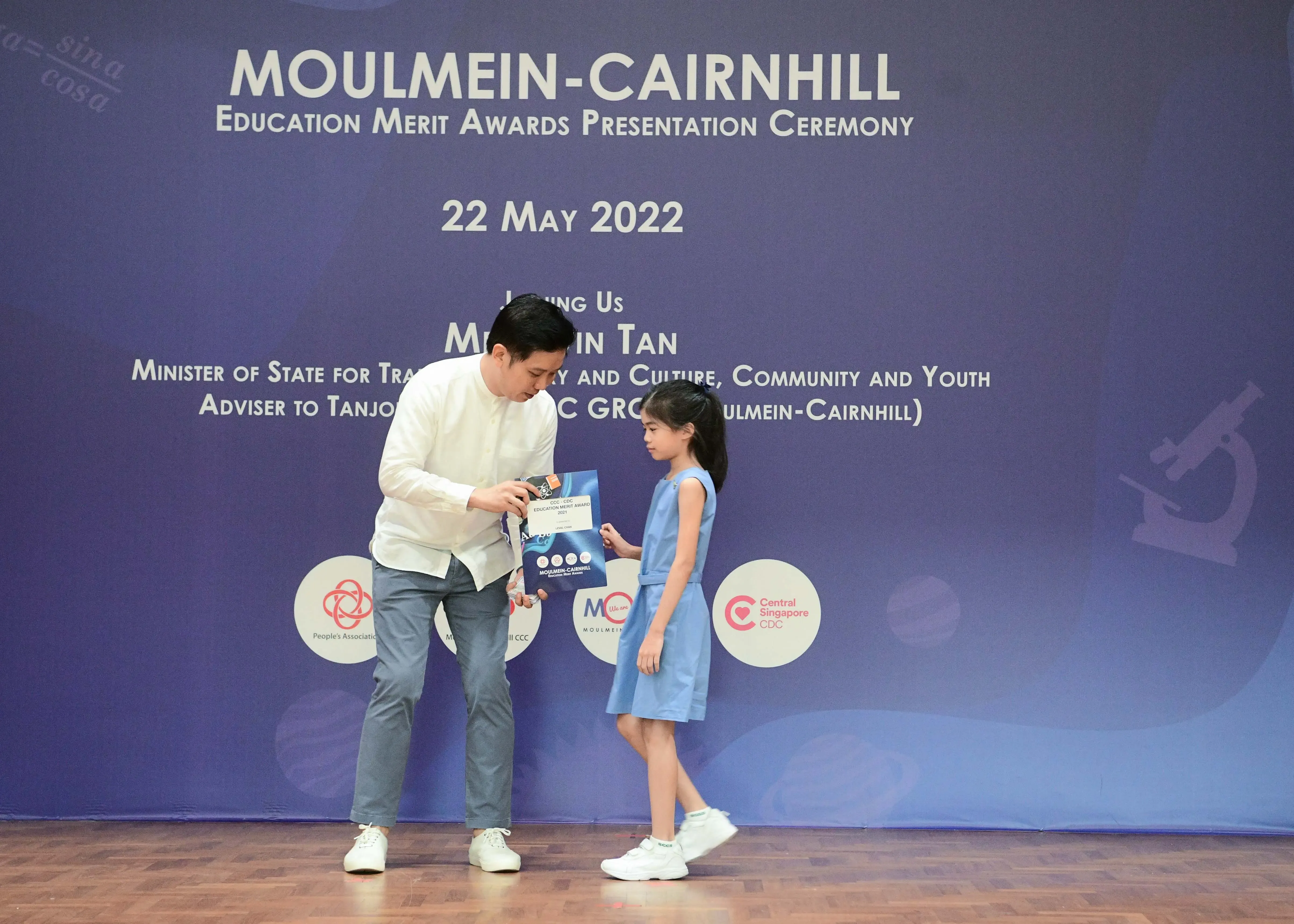 A man hands an award packet to a young girl on stage at the Moulmein-Cairnhill Education Merit Awards on 22 May 2022.