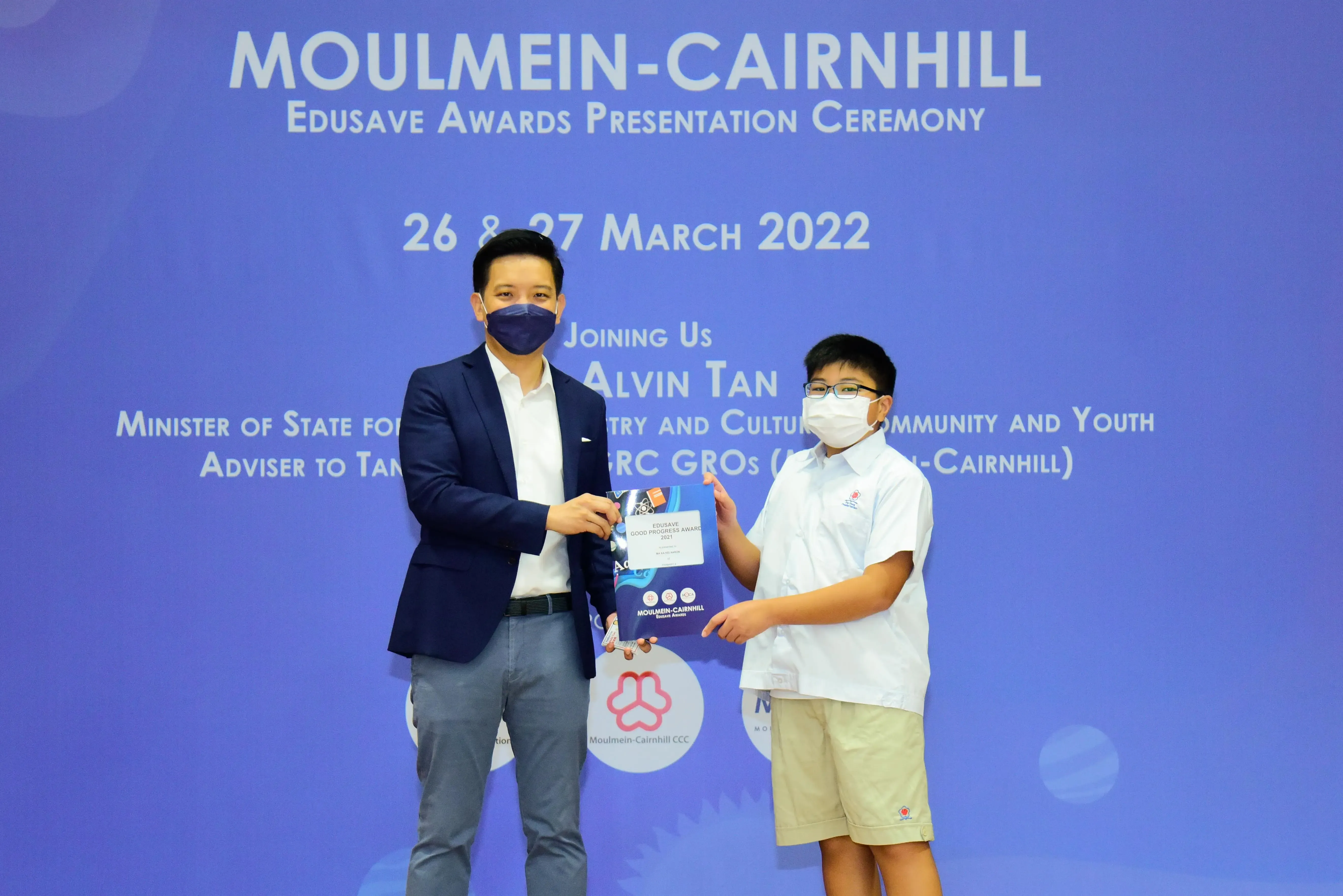 An adult in a blazer and a young student, both masked, hold an award at the Moulmein-Cairnhill Edusave ceremony.