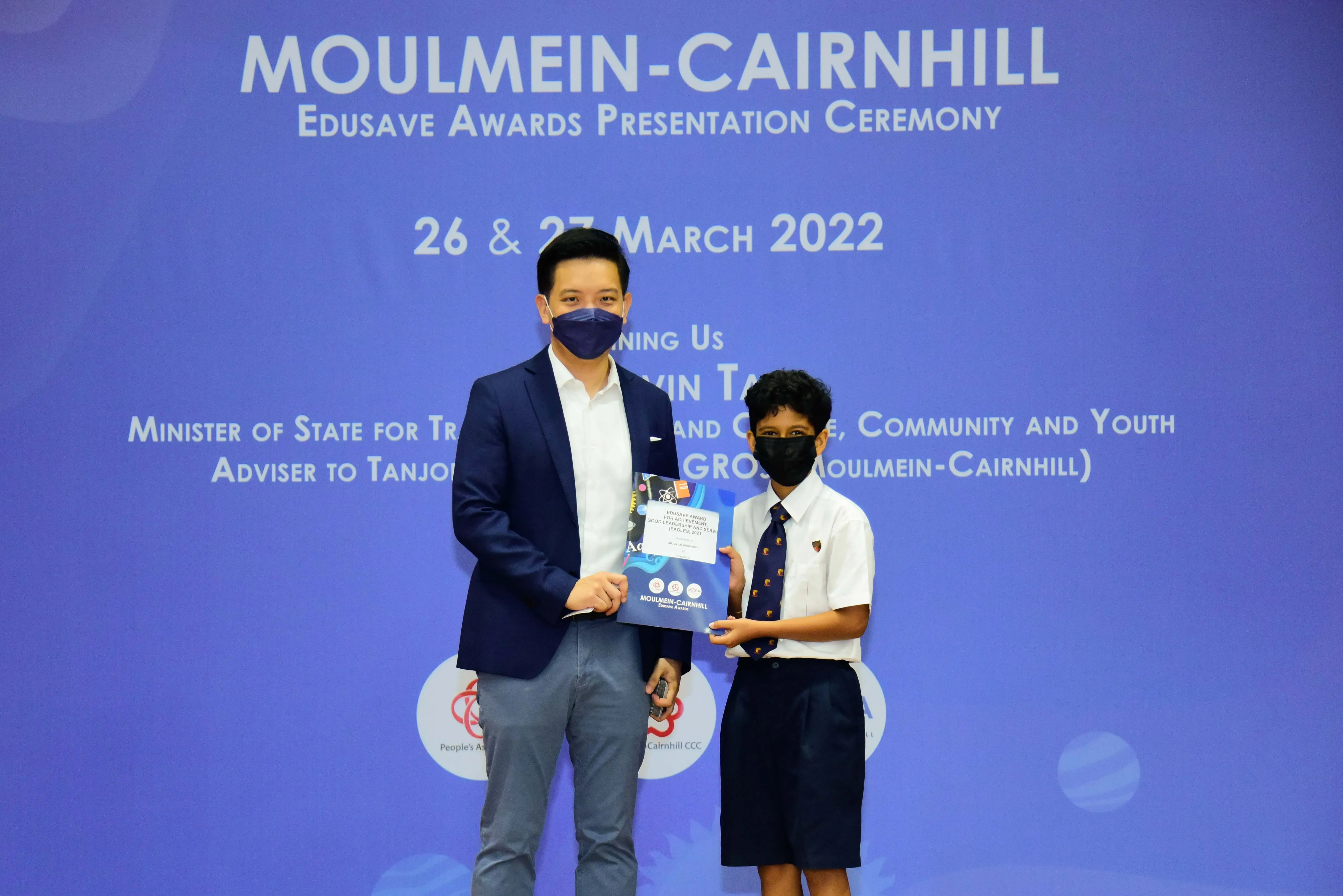 An adult and a masked schoolboy hold an award folder at the Moulmein-Cairnhill Edusave Awards Ceremony, March 2022.