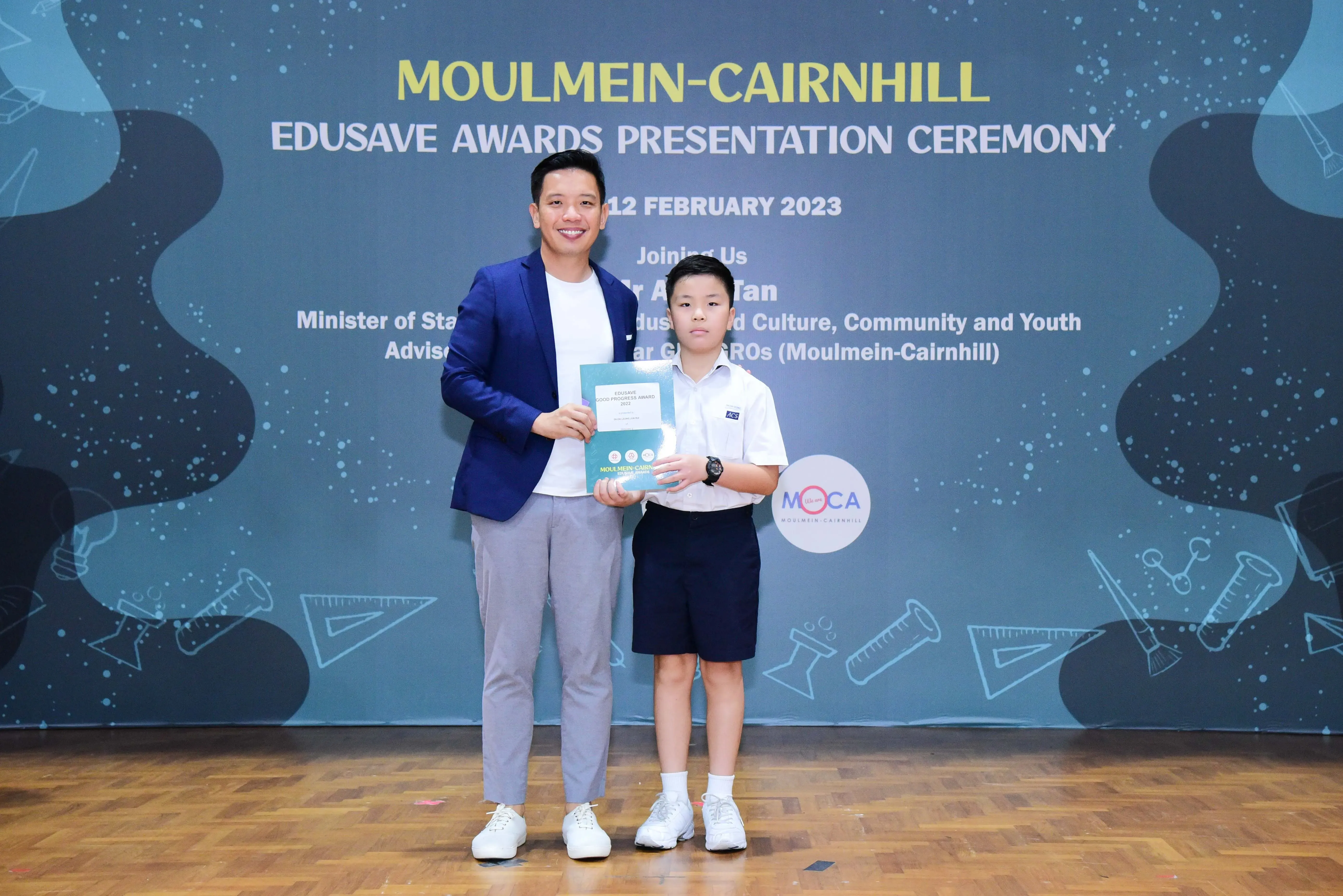 An adult in a blue blazer and a young student in uniform hold a certificate at the Moulmein-Cairnhill Edusave Awards 2023.