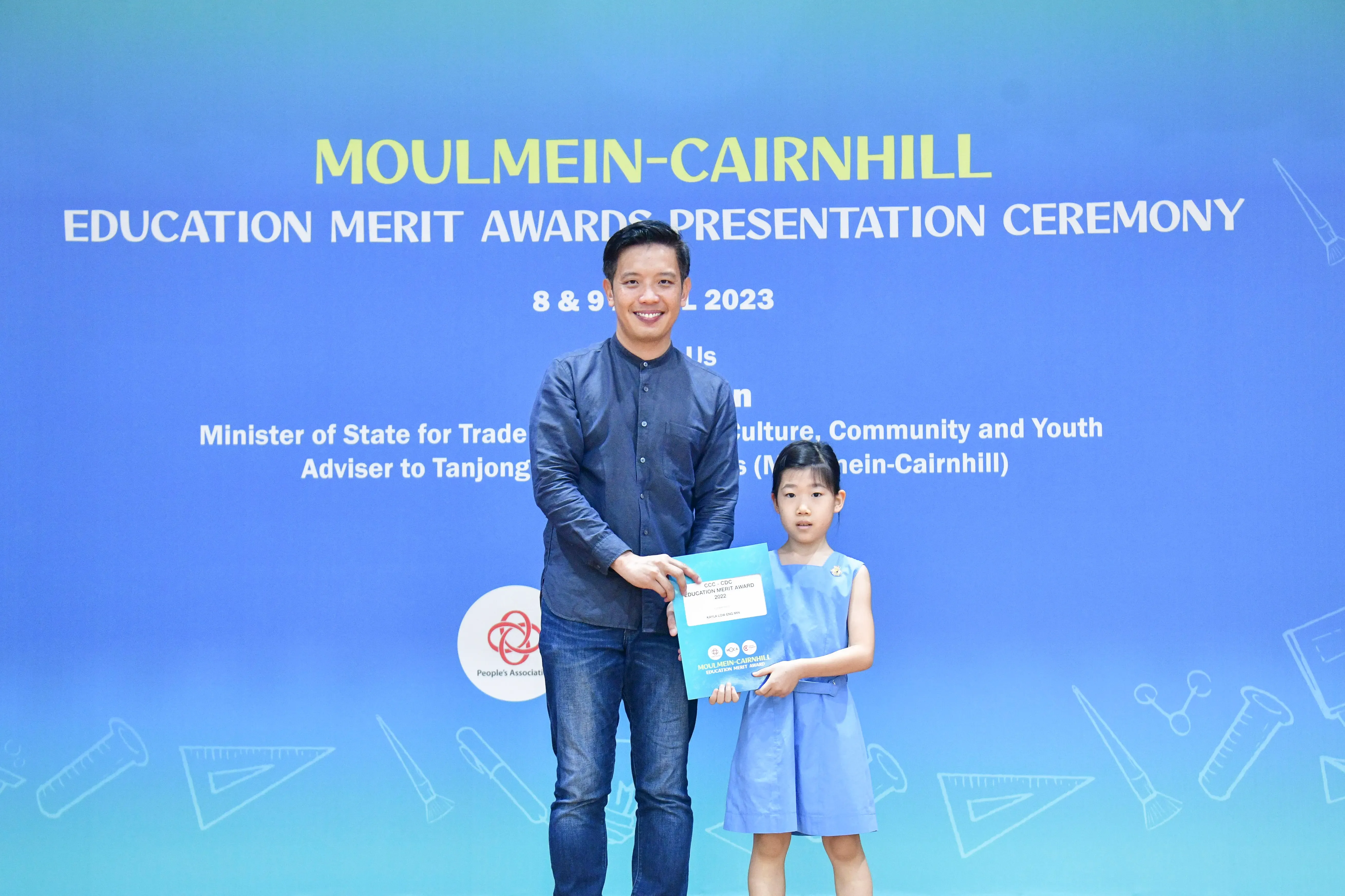 A man and a young girl hold a certificate together in front of a Moulmein-Cairnhill Education Merit Awards banner.