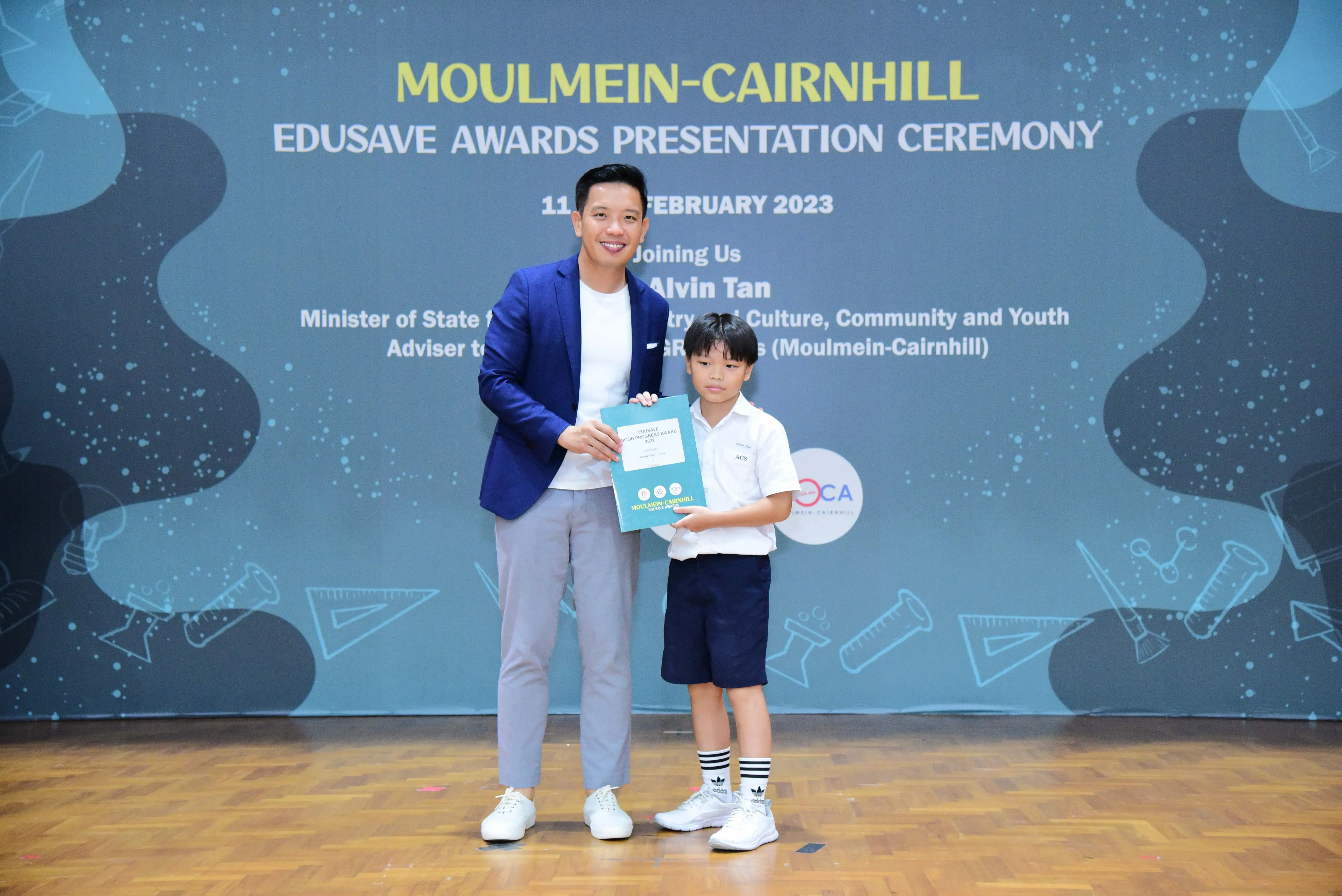 A man in a blue blazer and a young boy in school uniform hold a certificate at the Moulmein-Cairnhill Edusave Awards 2023.
