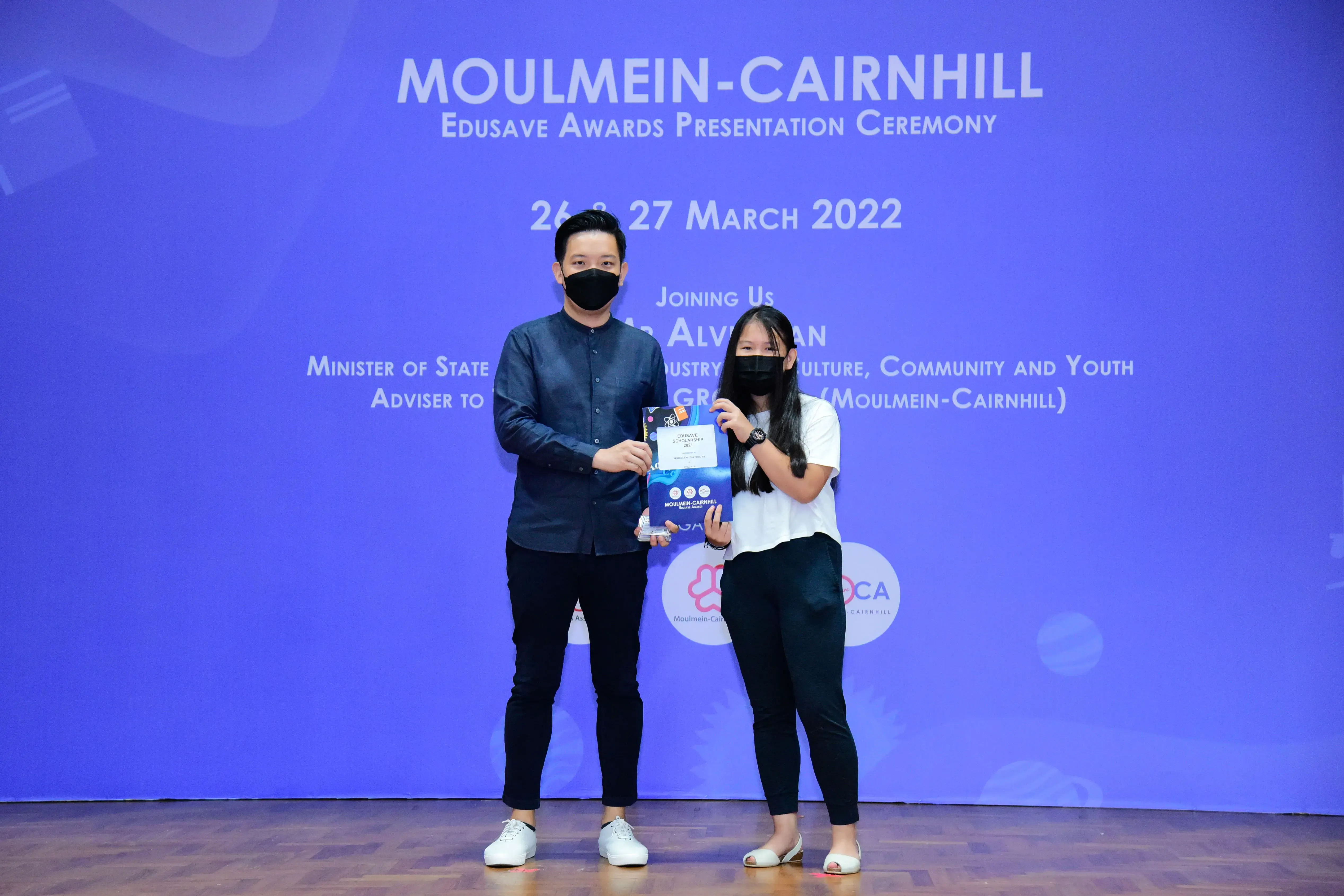 Two masked people hold an award certificate on stage at the Moulmein-Cairnhill Edusave Awards Ceremony, March 2022.