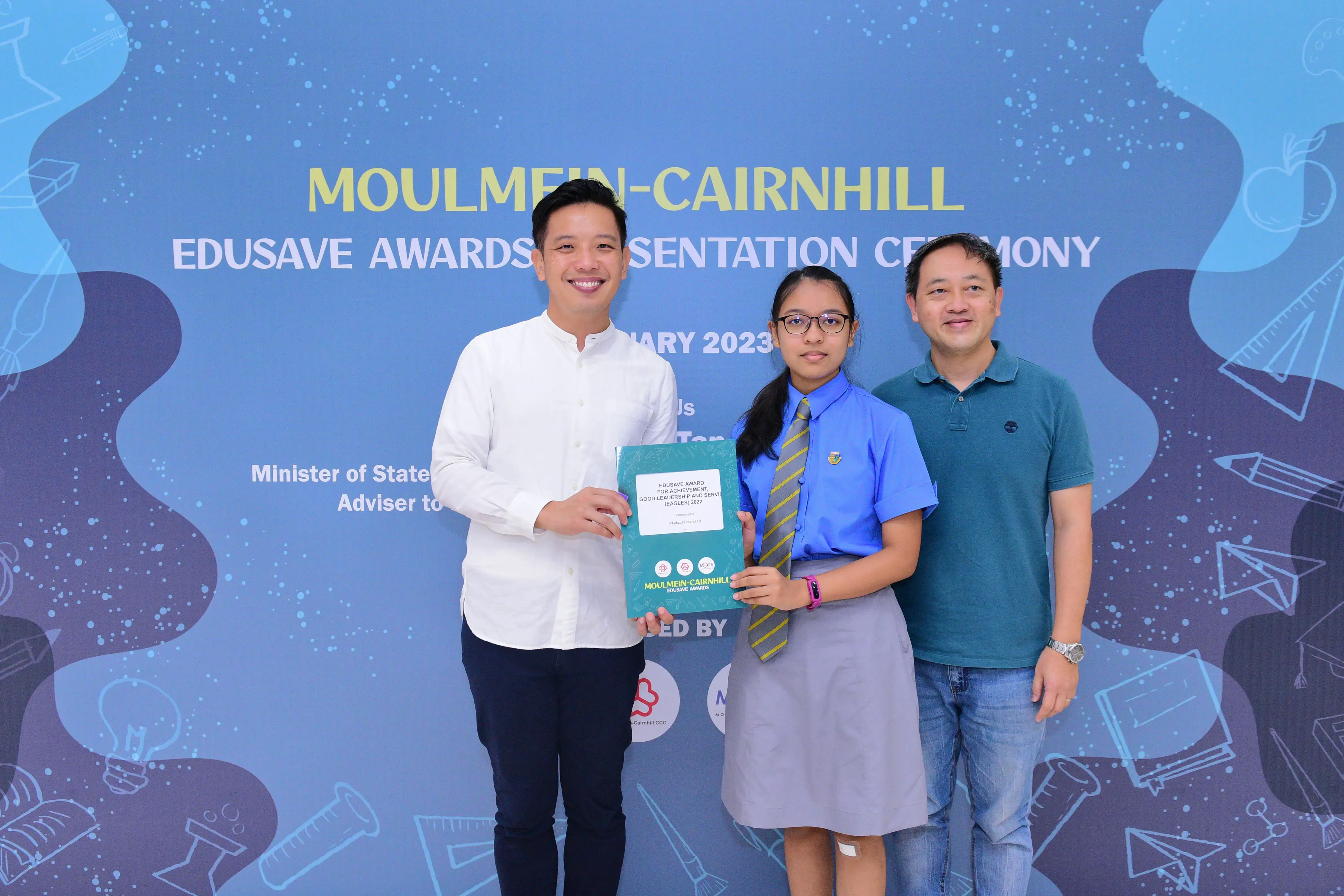 A student in school uniform holds a certificate flanked by two men at the Moulmein-Cairnhill Edusave Awards Ceremony 2023.