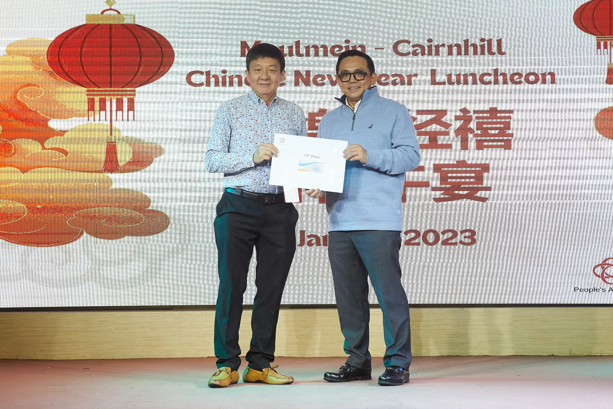 Two men hold a prize envelope on stage at the Moulmein-Cairnhill Chinese New Year Luncheon 2023.