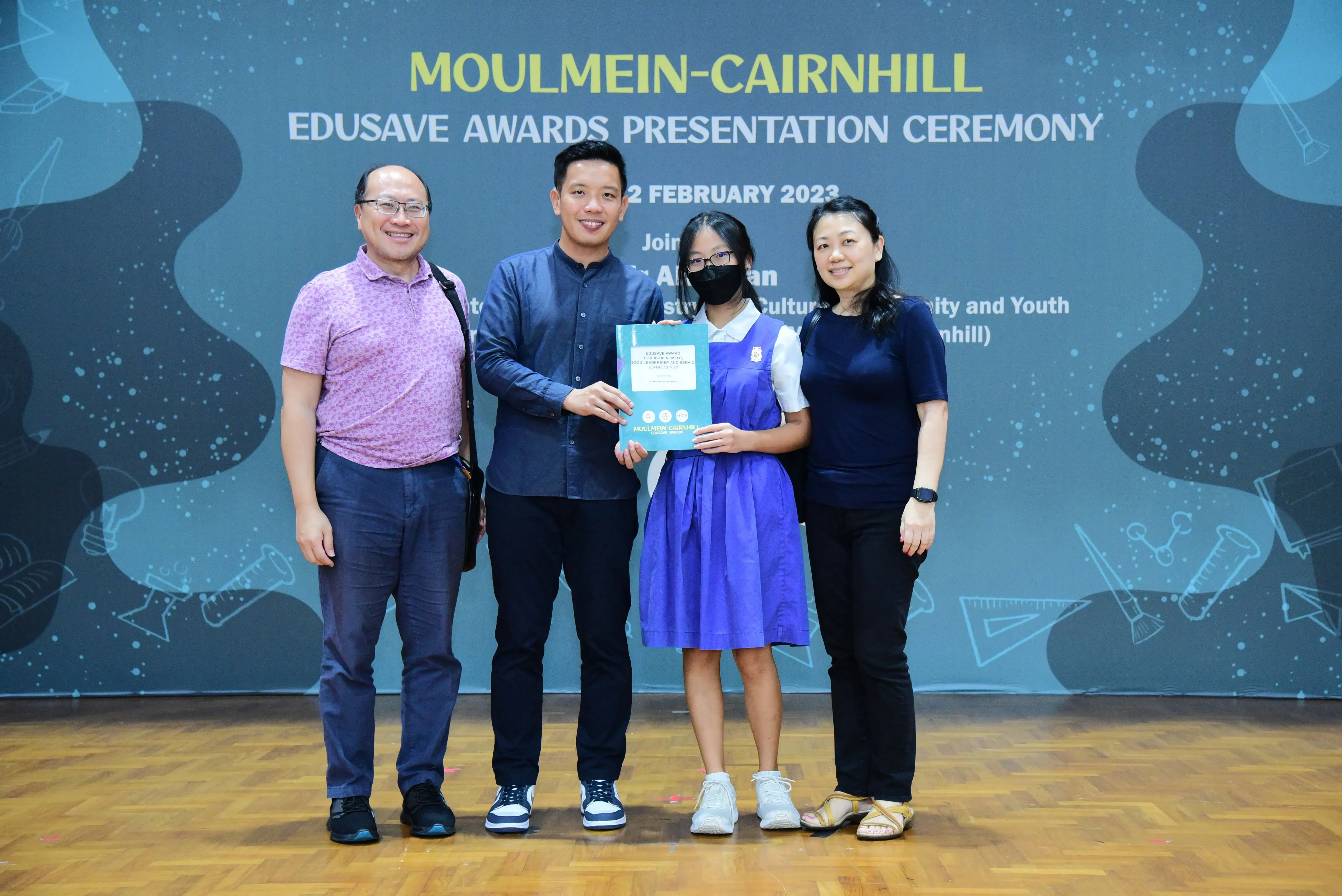 Four people pose at the Moulmein-Cairnhill Edusave Awards Presentation Ceremony, with a girl holding a certificate.