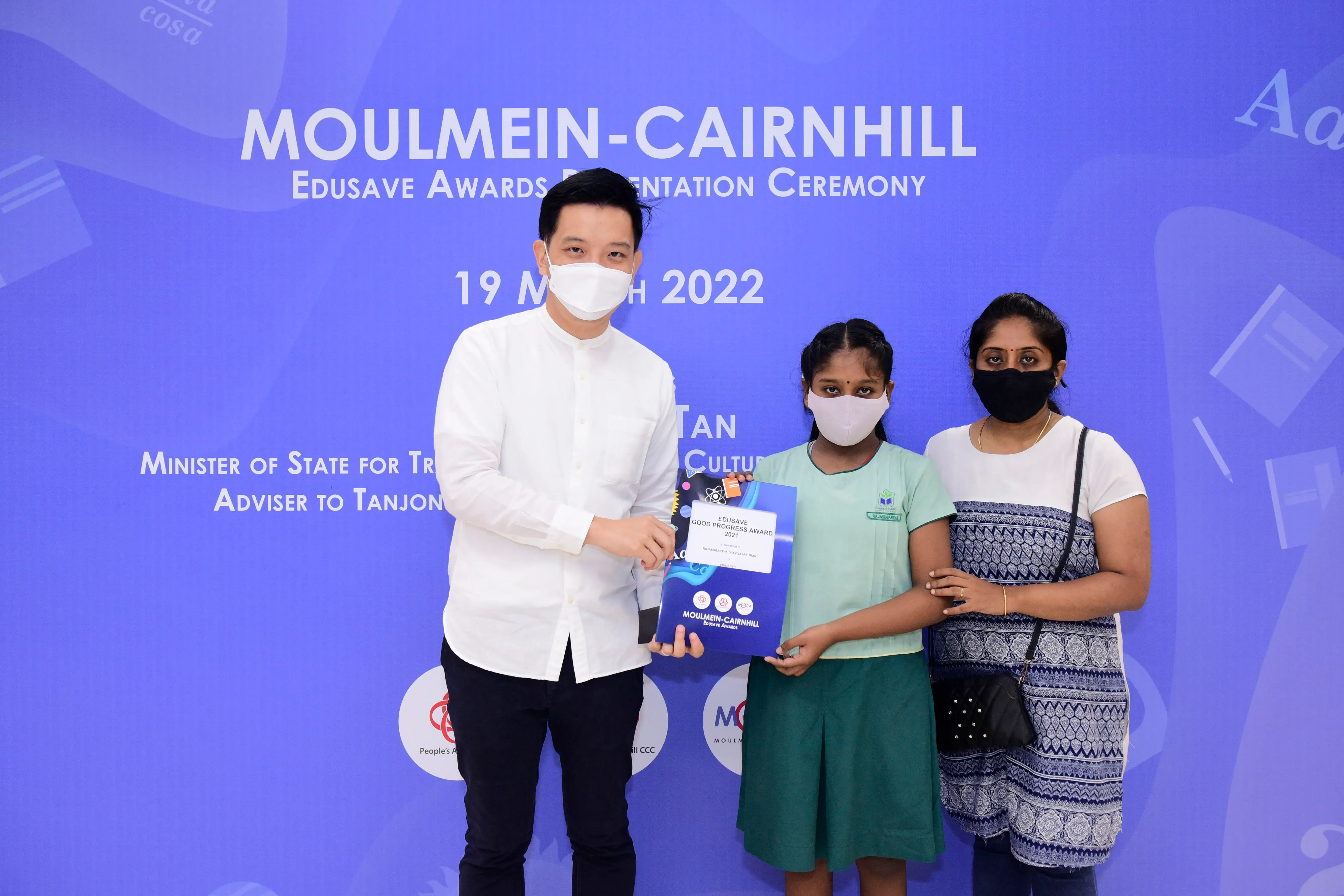 Three masked people pose at the Moulmein-Cairnhill Edusave Awards Presentation Ceremony, holding an award folder.