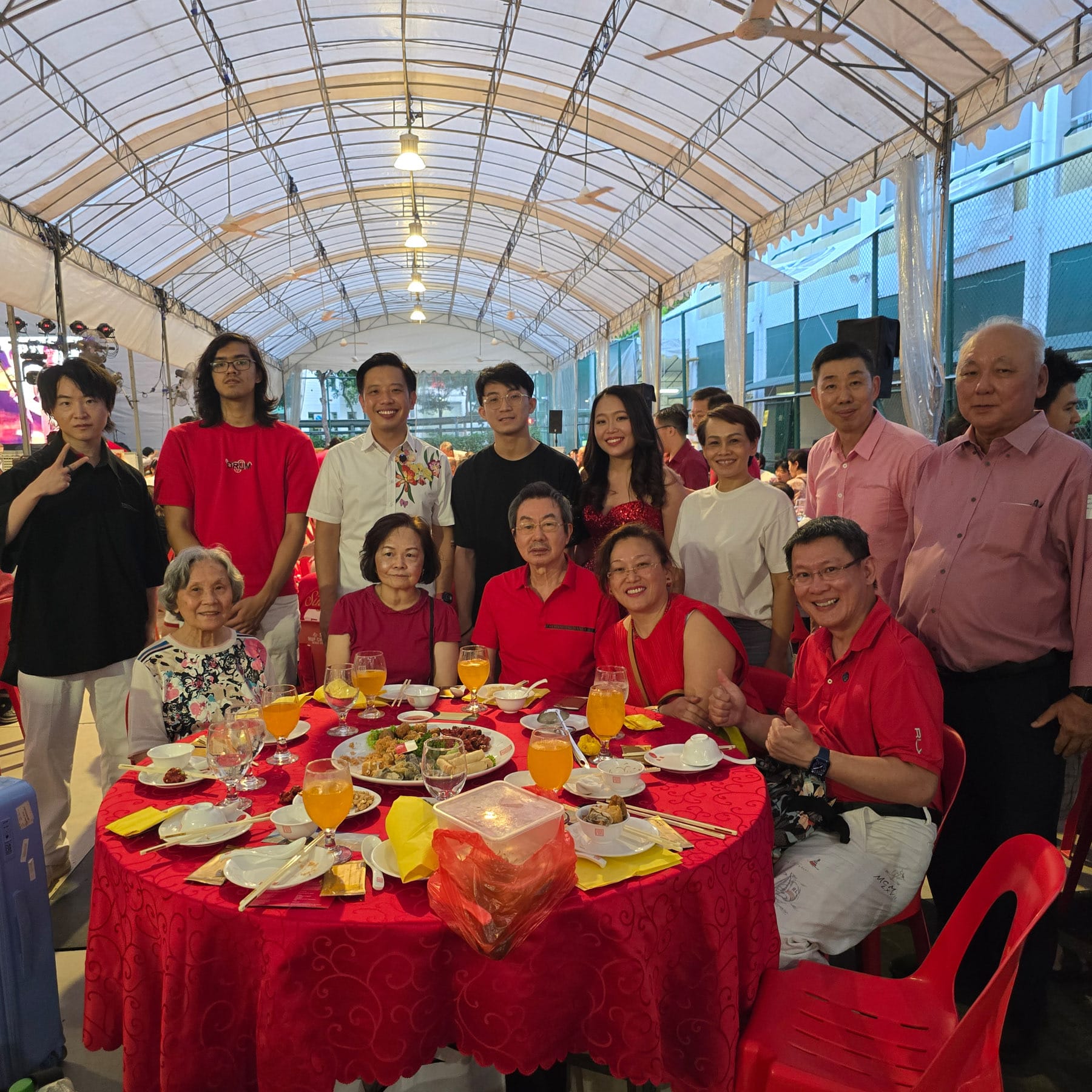 A large group of people in red outfits pose around a red banquet table with food and orange juice under a tent.