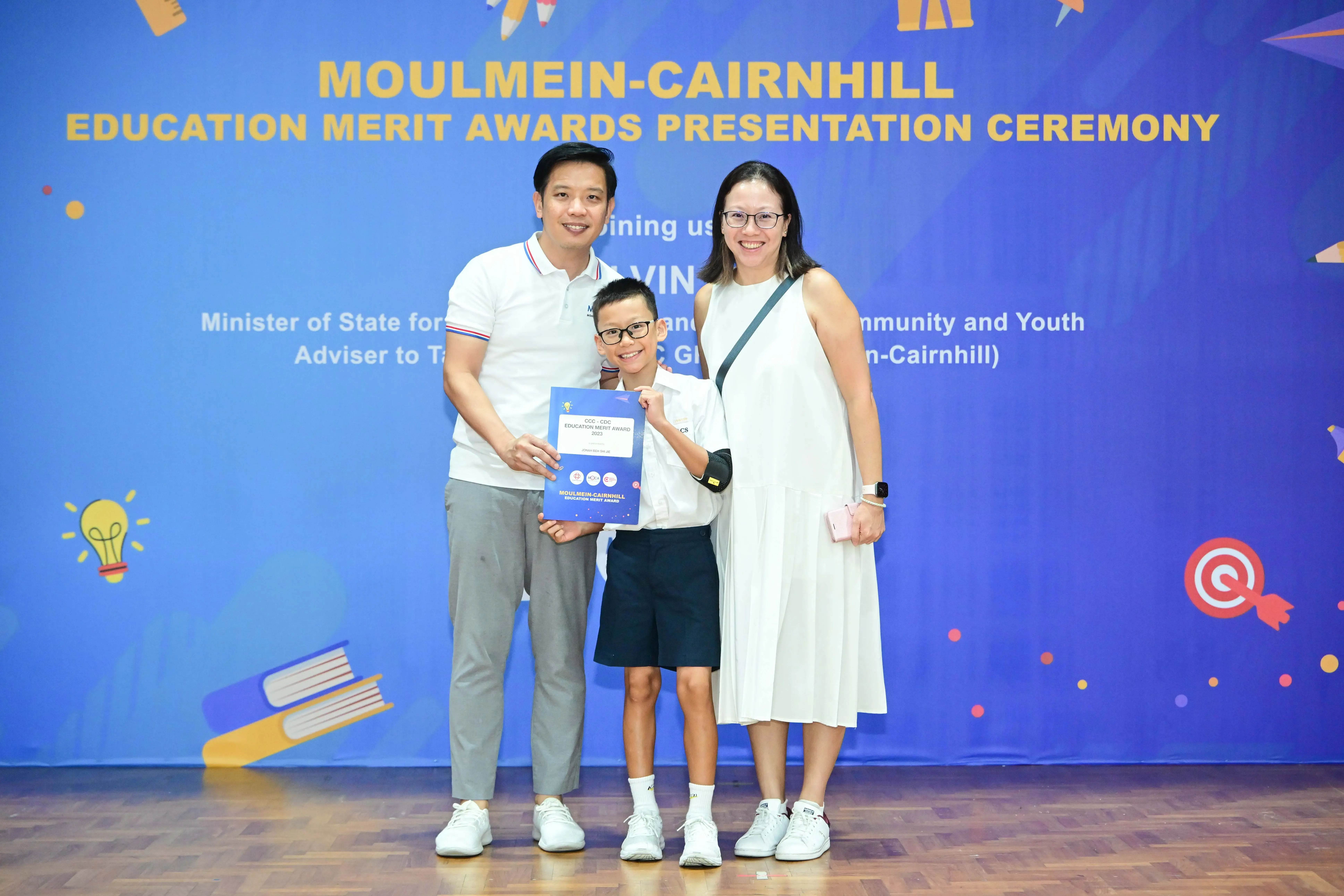 A boy holding a merit award certificate stands between two smiling adults at the Moulmein-Cairnhill Education Merit Awards ceremony.
