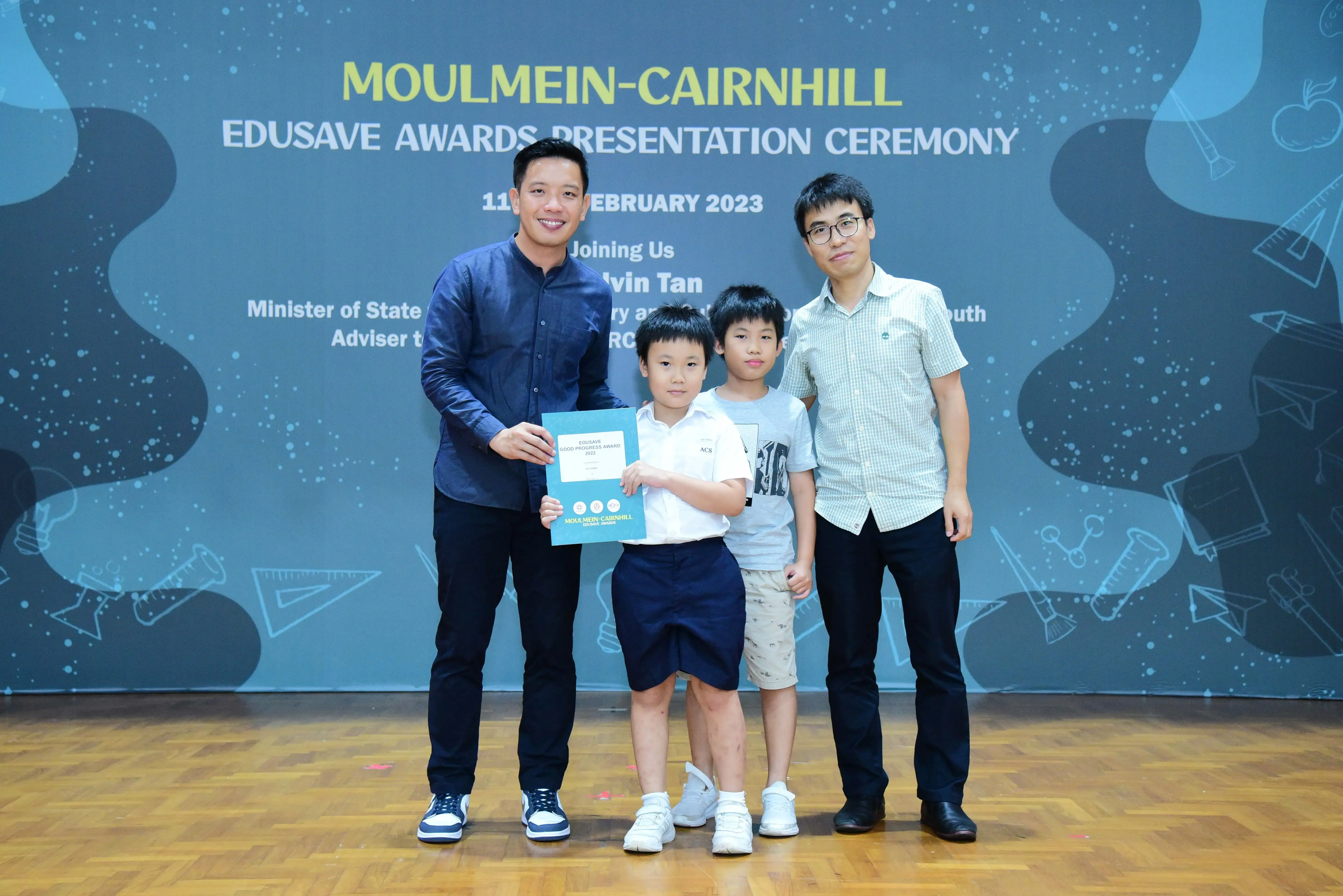 Two men and two young boys pose with a certificate at the Moulmein-Cairnhill Edusave Awards Ceremony, February 2023.