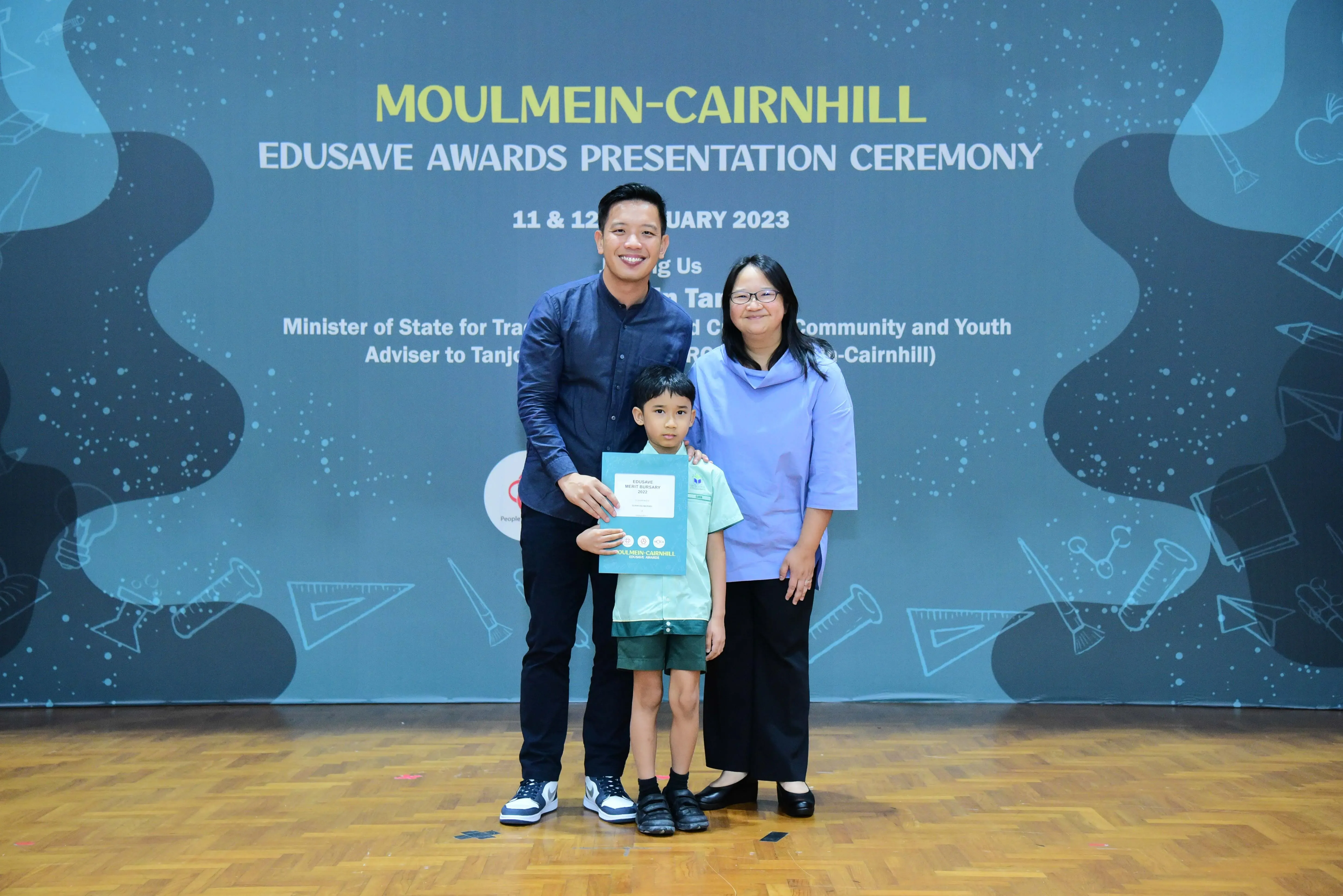 Two adults and a young boy holding a certificate pose at the Moulmein-Cairnhill Edusave Awards Ceremony 2023.