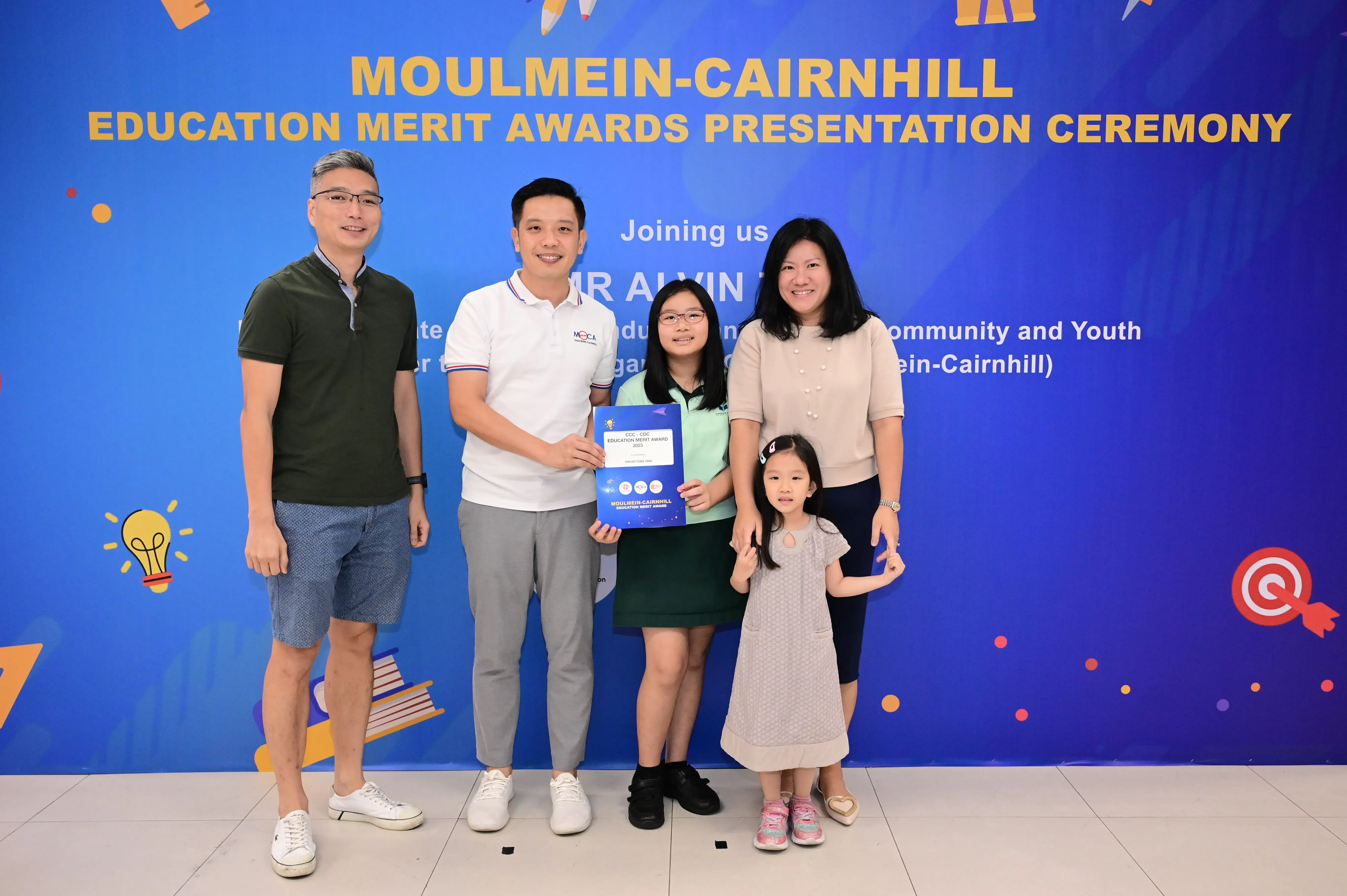 Five people pose with a certificate at the Moulmein-Cairnhill Education Merit Awards Presentation Ceremony backdrop.