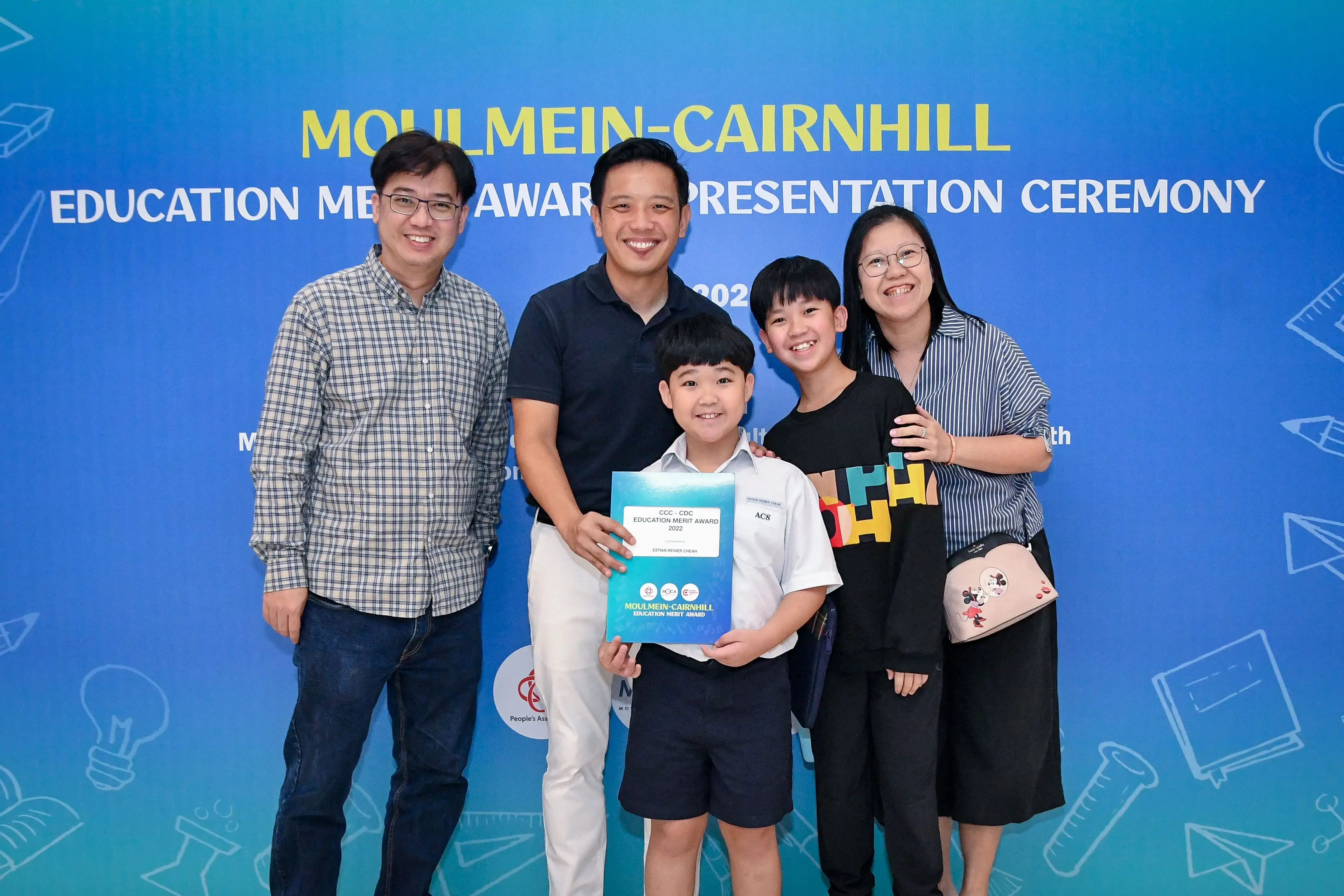 Five smiling people pose at the Moulmein-Cairnhill Education Merit Award Presentation Ceremony, with a boy holding a certificate.